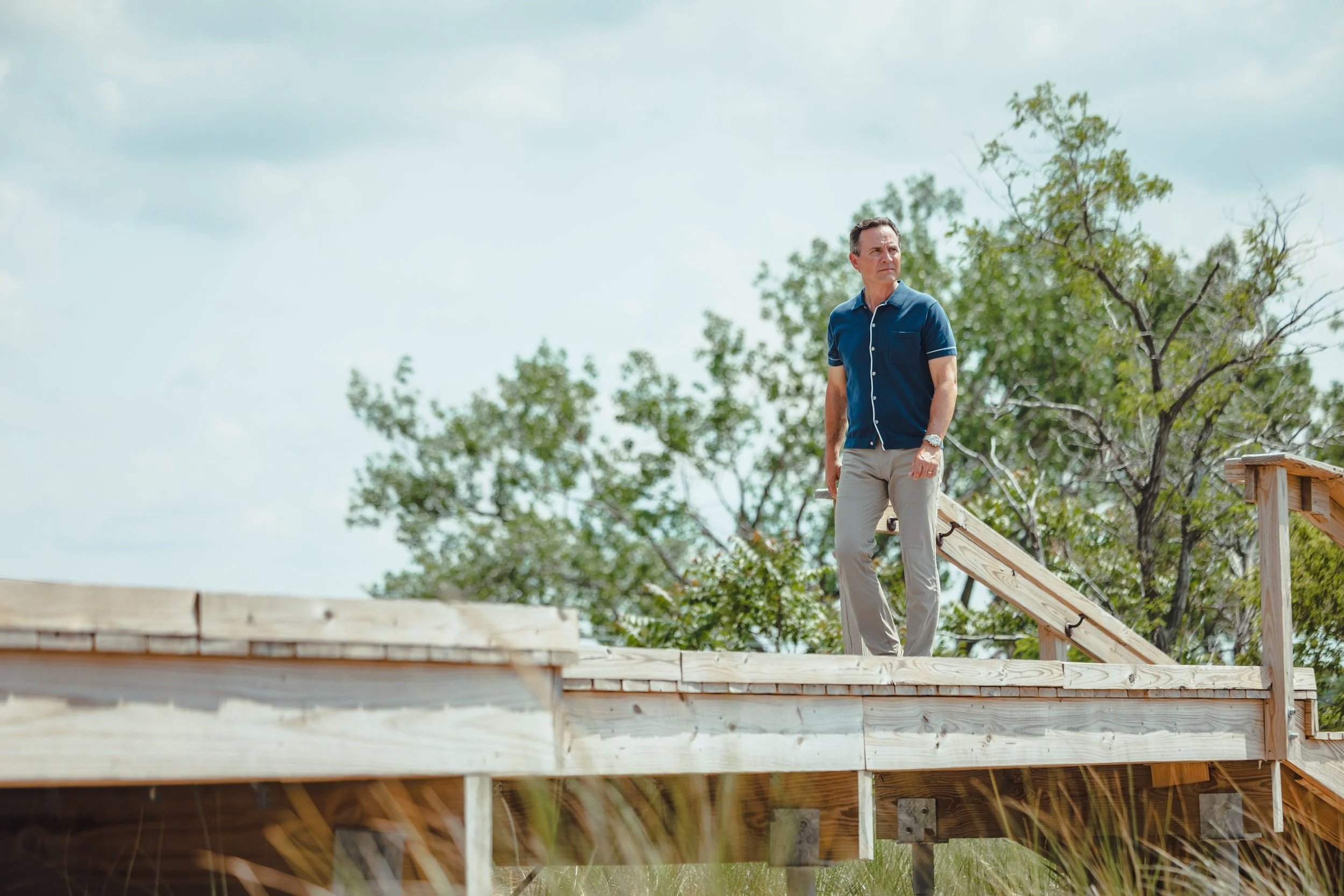 A man standing on a wooden deck under a cloudy sky surrounded by trees.