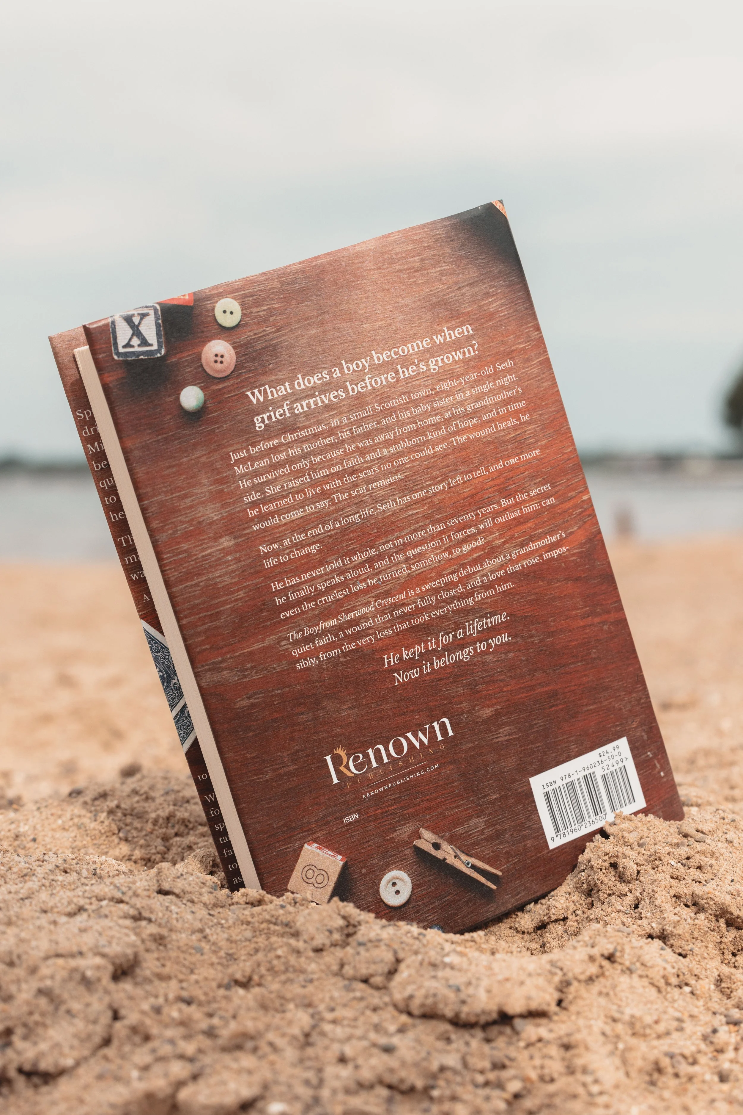 The back cover of the book on a sandy beach. The book is partly buried in the sand with a cloudy sky in the background. The back cover of the book features text, buttons, and game pieces, including dice, buttons, and game tokens.
