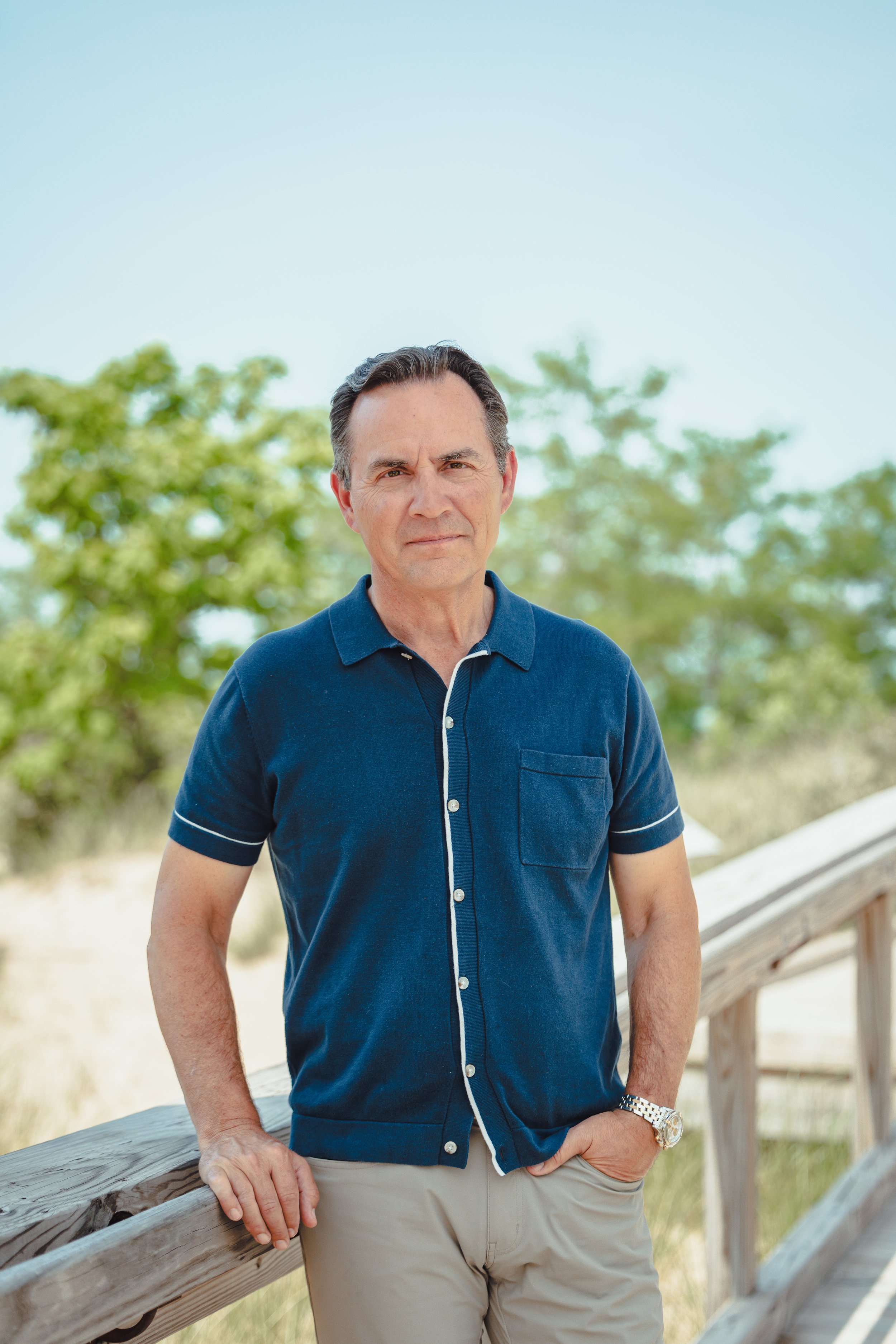 A middle-aged man standing outdoors on a wooden railing bridge, wearing a blue short-sleeved shirt and beige pants, with a background of trees and a bright, clear sky.