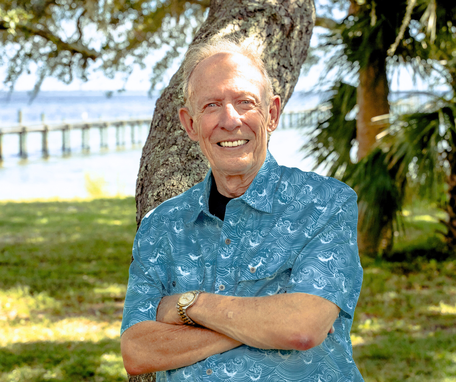 An elderly man standing outdoors near a tree with arms crossed, smiling, with water and a dock in the background.