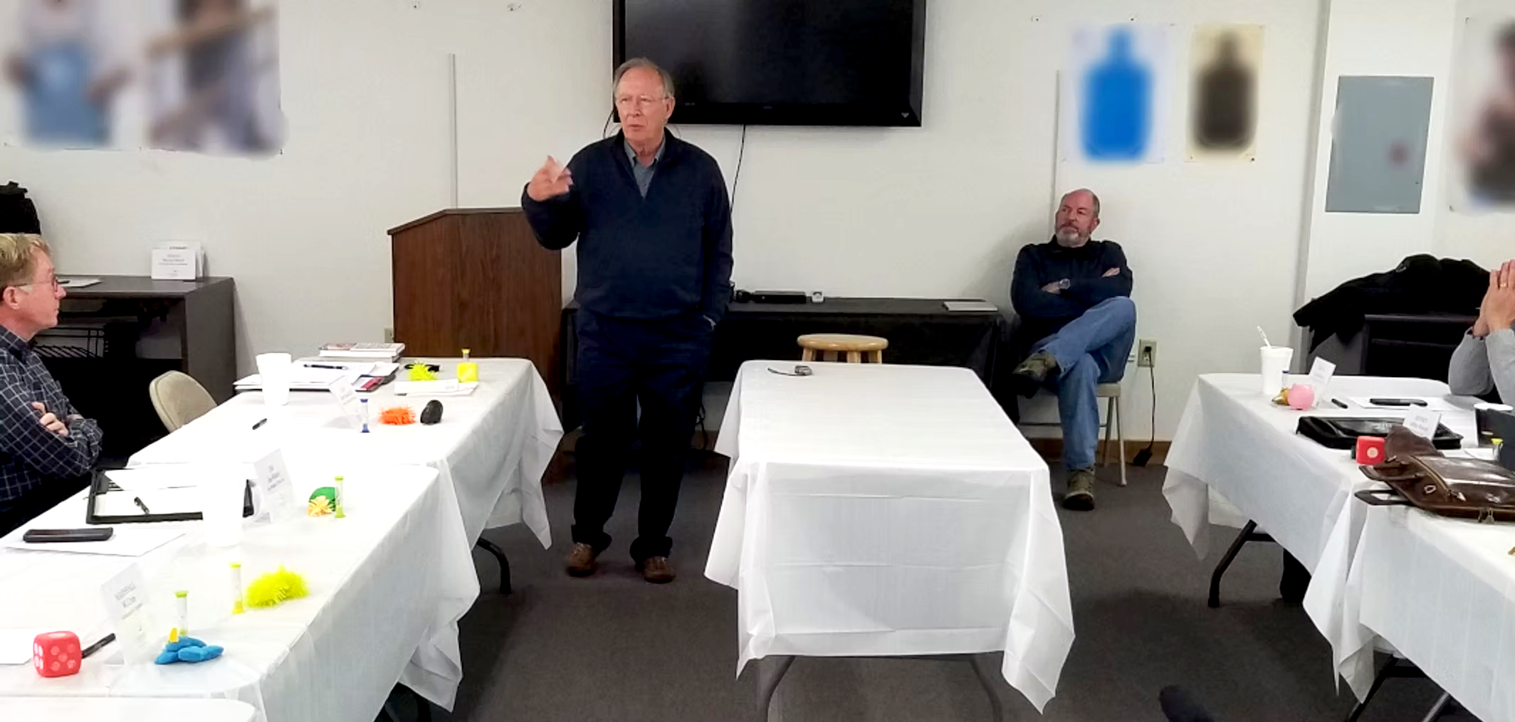 Art Kimbrough standing at the front of a room giving a presentation to seated attendees, with a large TV screen behind him and chairs with tables on either side.