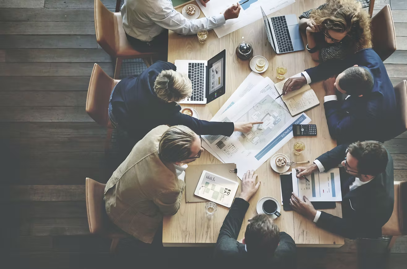 Business meeting with seven people gathered around a wooden table, reviewing architectural plans, using laptops, tablets, and papers, with drinks and snacks on the table.