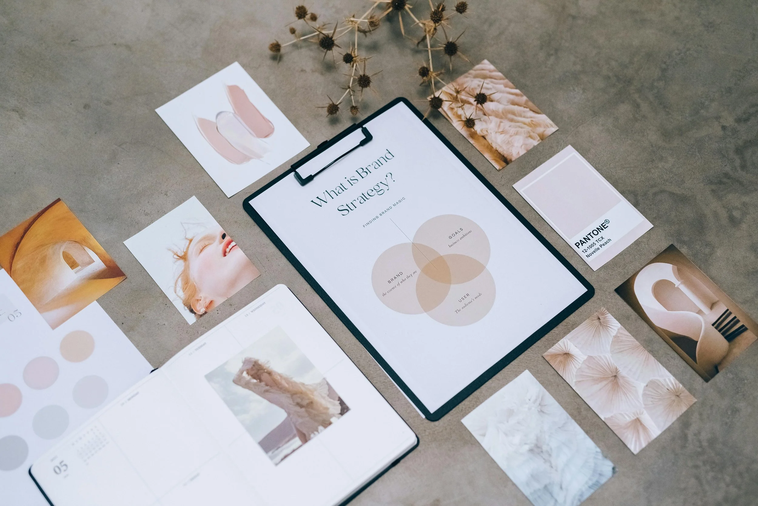 Flat lay of design mood board with sample photos, color swatches, and a tablet displaying a presentation on brand strategy, surrounded by dried flowers, a notebook, and sample images