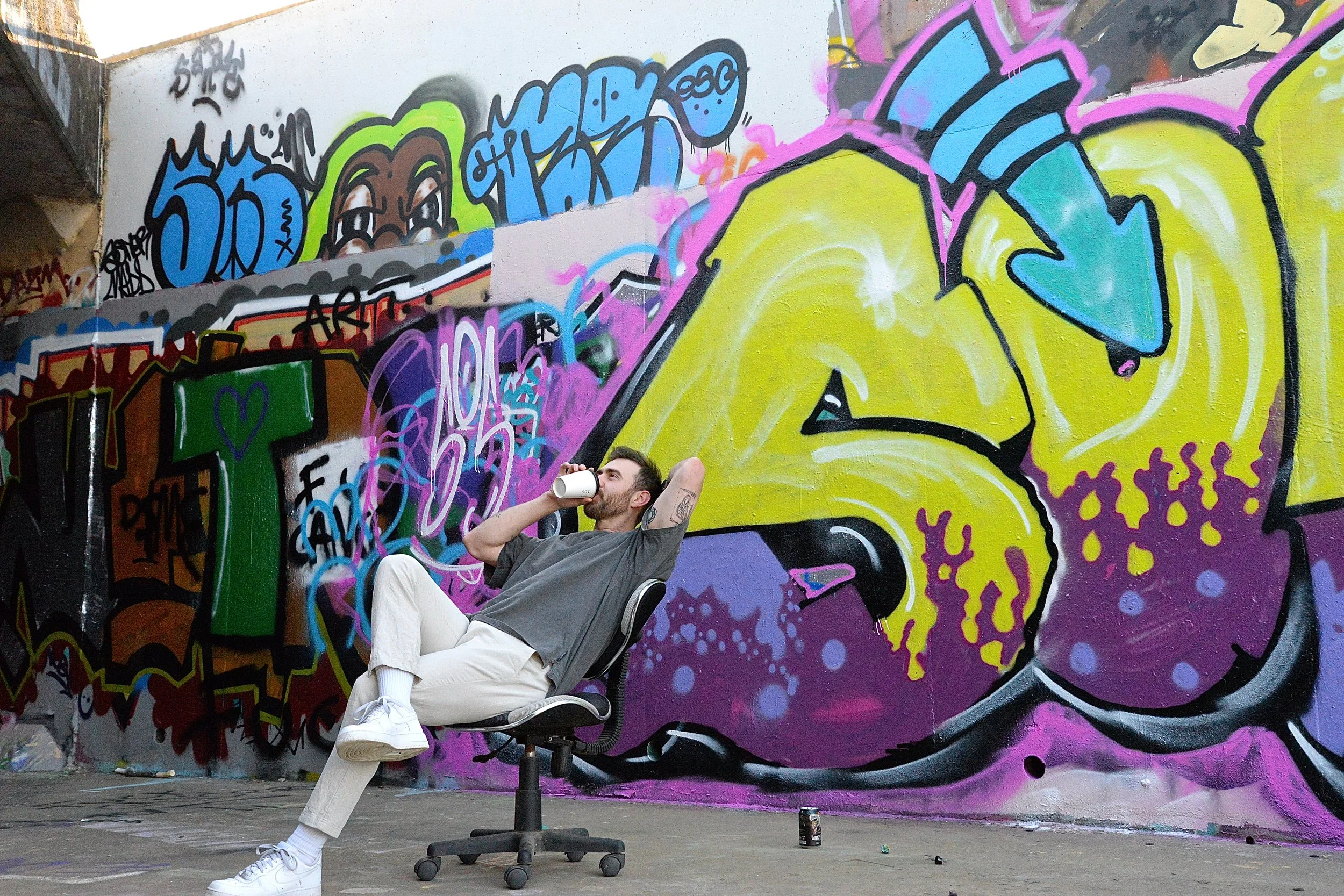A man sitting on an office chair with his legs crossed, drinking from a white cup, in front of a colorful graffiti wall. The graffiti features various vibrant colors and abstract designs.