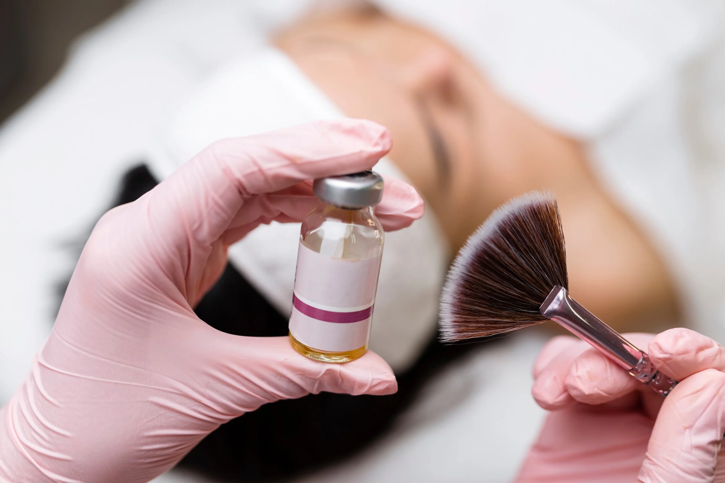 A beautician in pink gloves holding a small bottle of oil or serum and a fan brush, preparing to apply a skincare treatment to a woman's face lying back with her eyes closed.