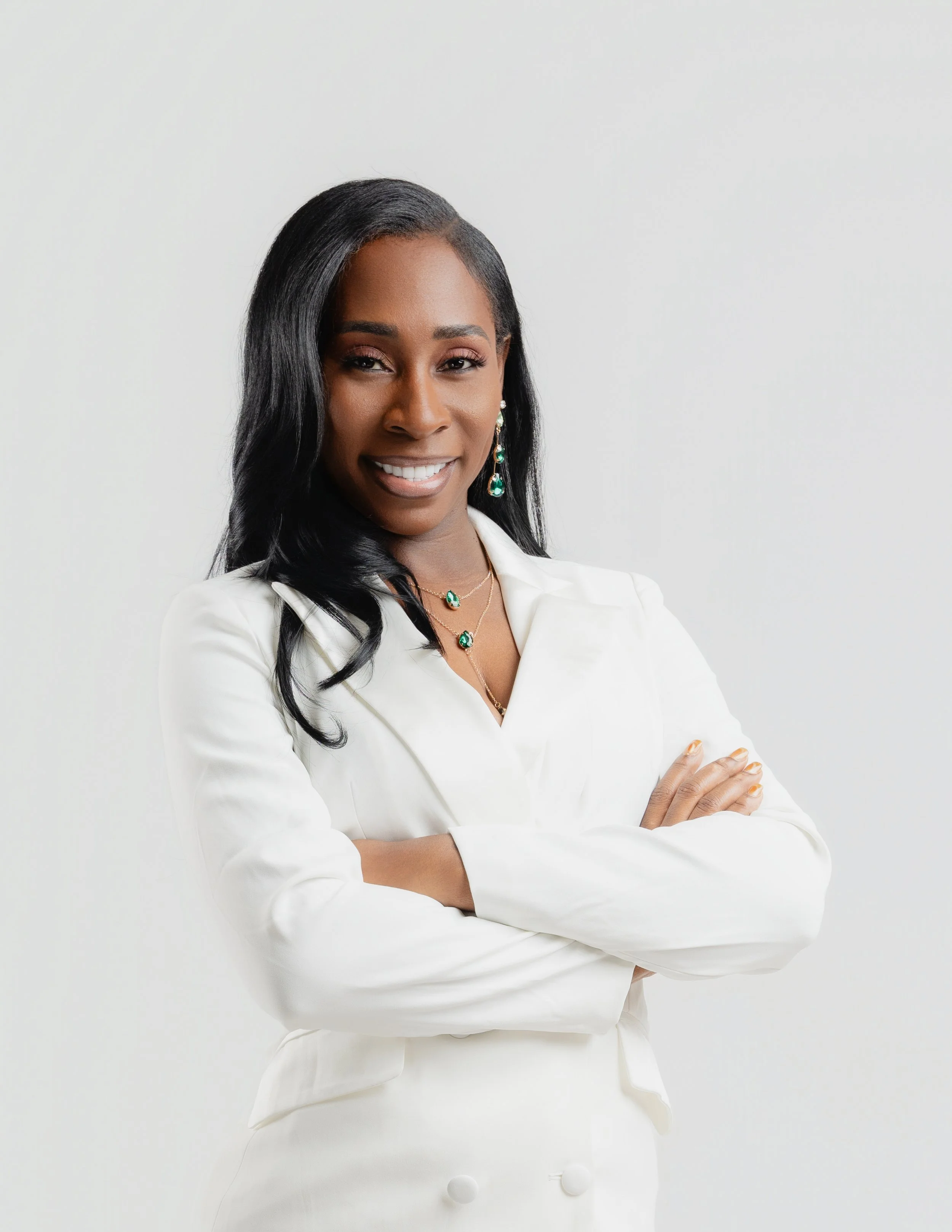 A confident African American woman with long black hair, dressed in a white blazer, smiling with arms crossed, standing against a plain white background.