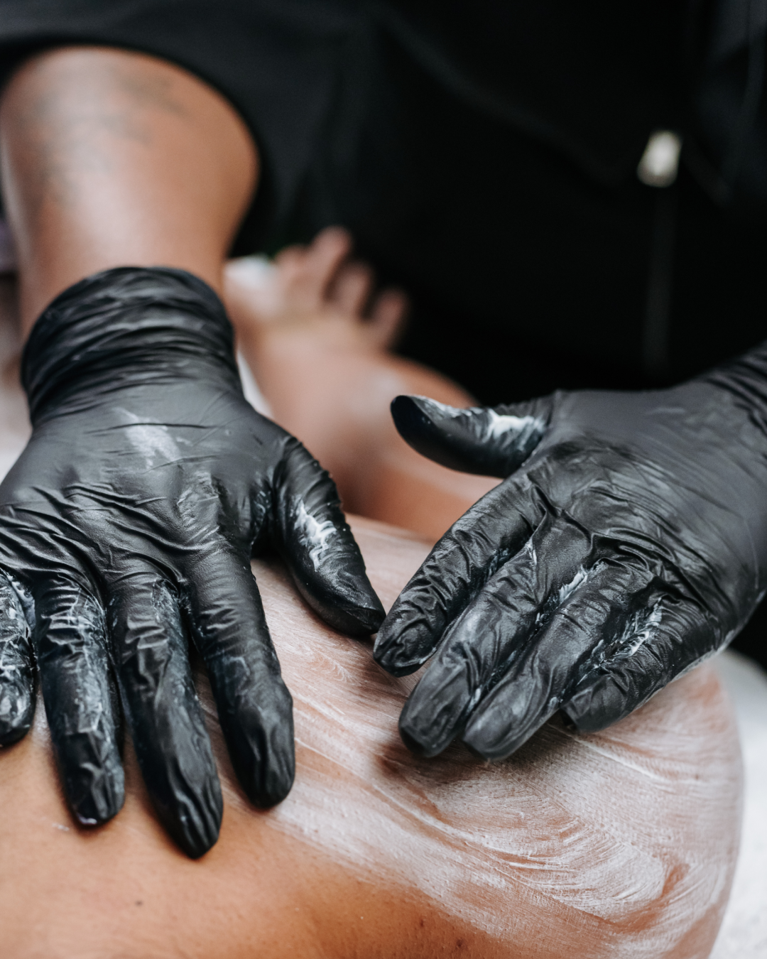Close-up of a person receiving a massage, with massage therapist wearing black gloves applying lotion or oil on the person's back.