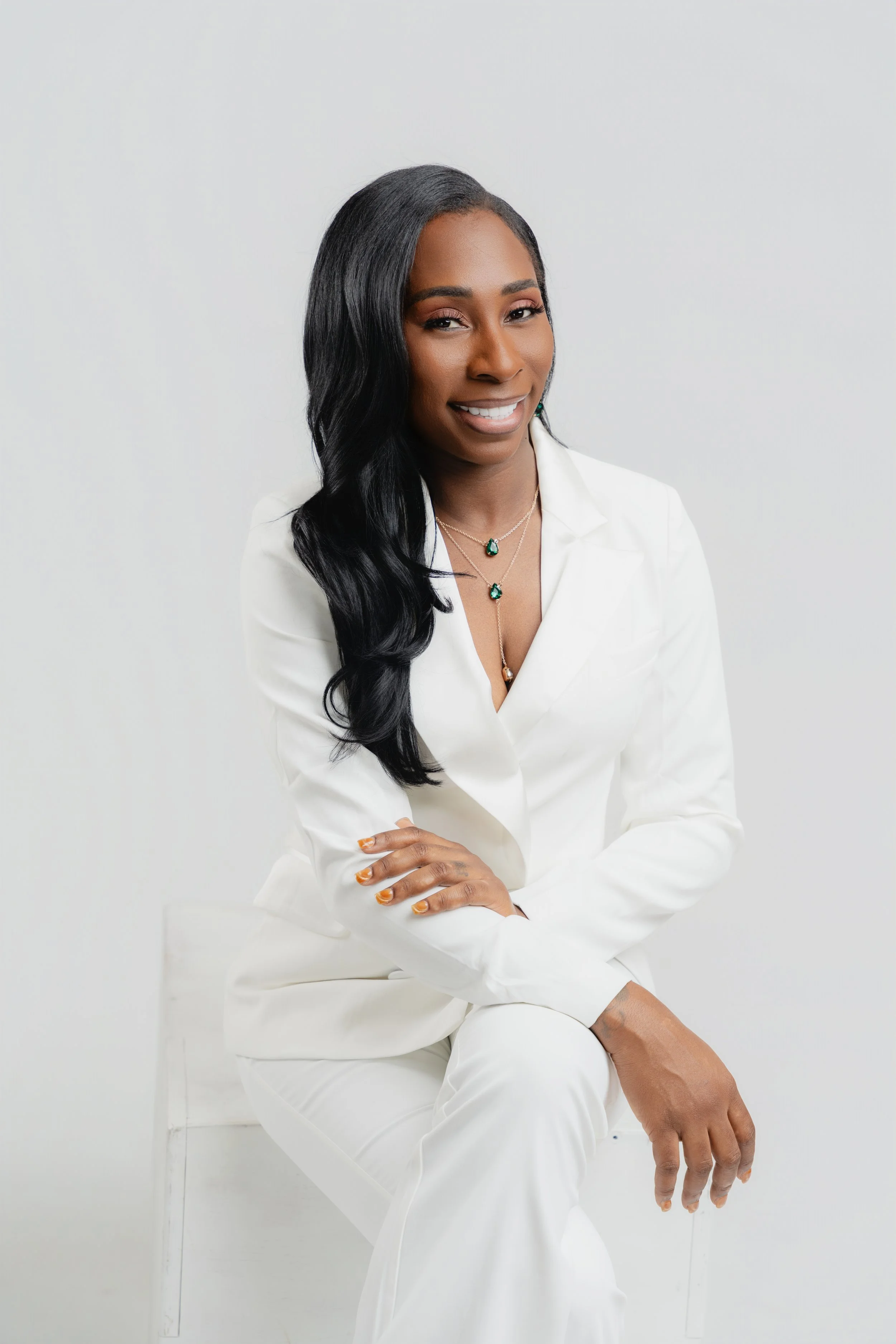 A woman with long black hair wearing a white blazer and matching white pants, sitting with one arm crossed over her knee, smiling against a white background, accessorized with layered necklaces with green gemstones.
