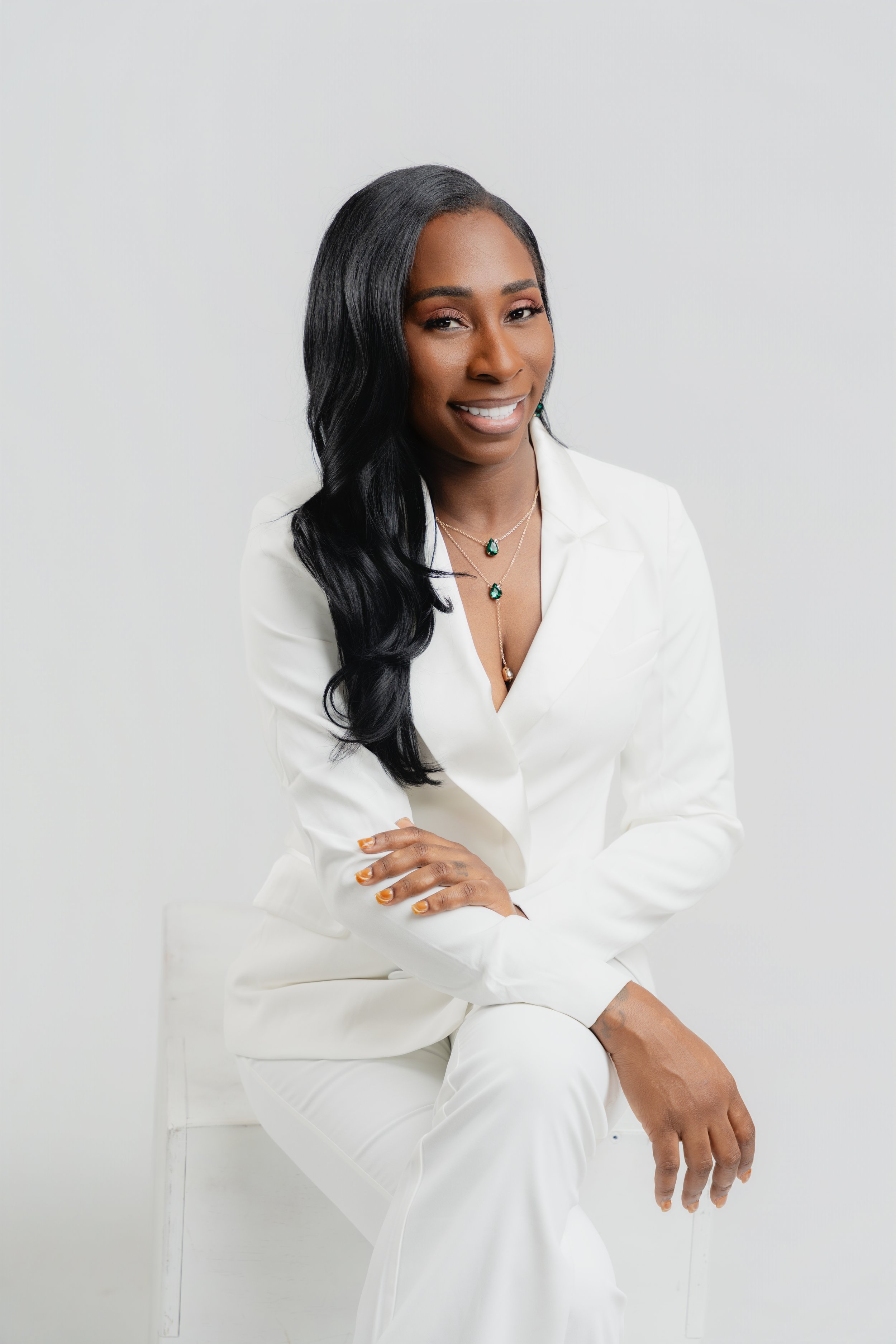 A woman with long black hair styled in loose waves, wearing a white suit, sitting on a white chair against a plain white background, smiling and looking at the camera.