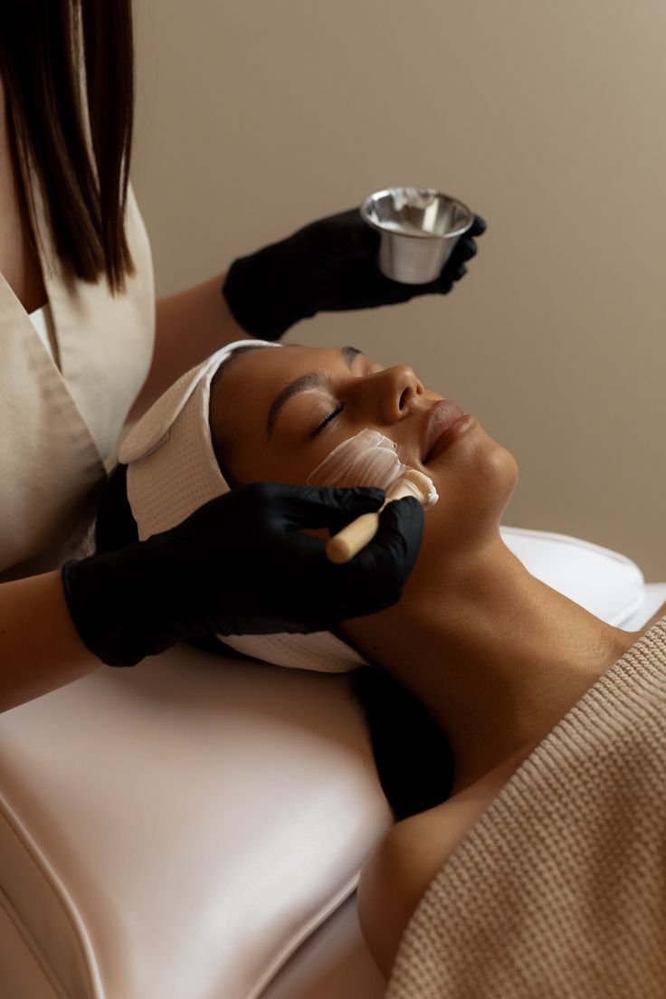 A woman receiving a facial treatment at a spa, with her eyes closed and her face relaxed, while a skincare professional applies a product using a brush.