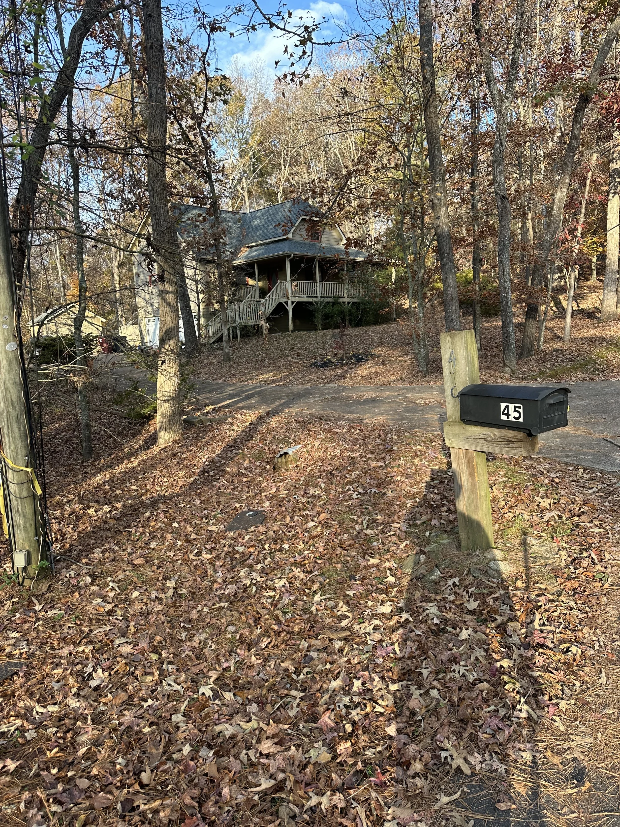 A house on a hill surrounded by trees with autumn leaves. A black mailbox with the number 45 is mounted on a wooden post near a driveway covered with fallen leaves.
Before