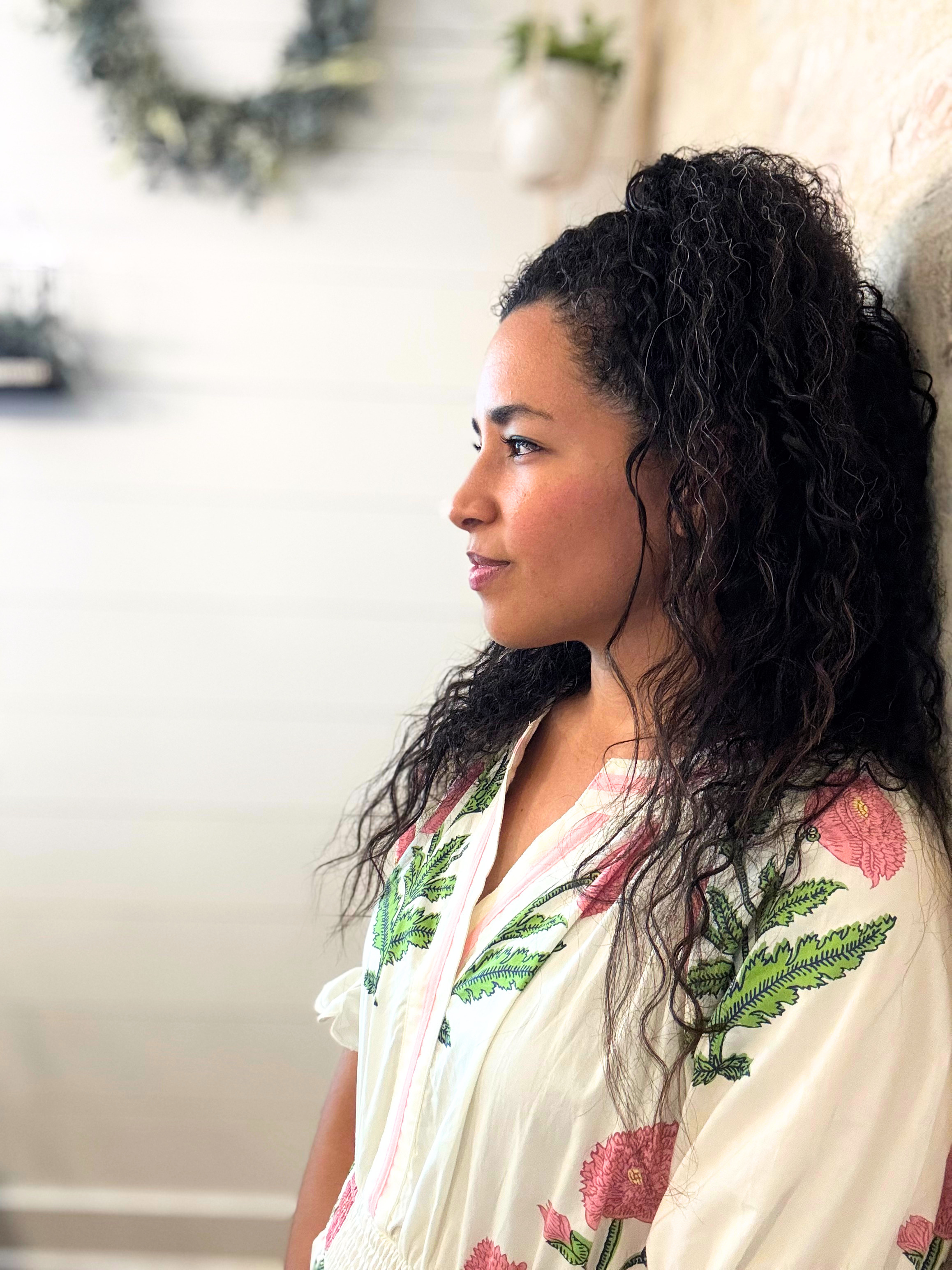 A young woman with curly dark hair and medium skin tone, looking to the right, standing against a light-colored wall. She is wearing a cream-colored kimono with pink flower and green leaf patterns.