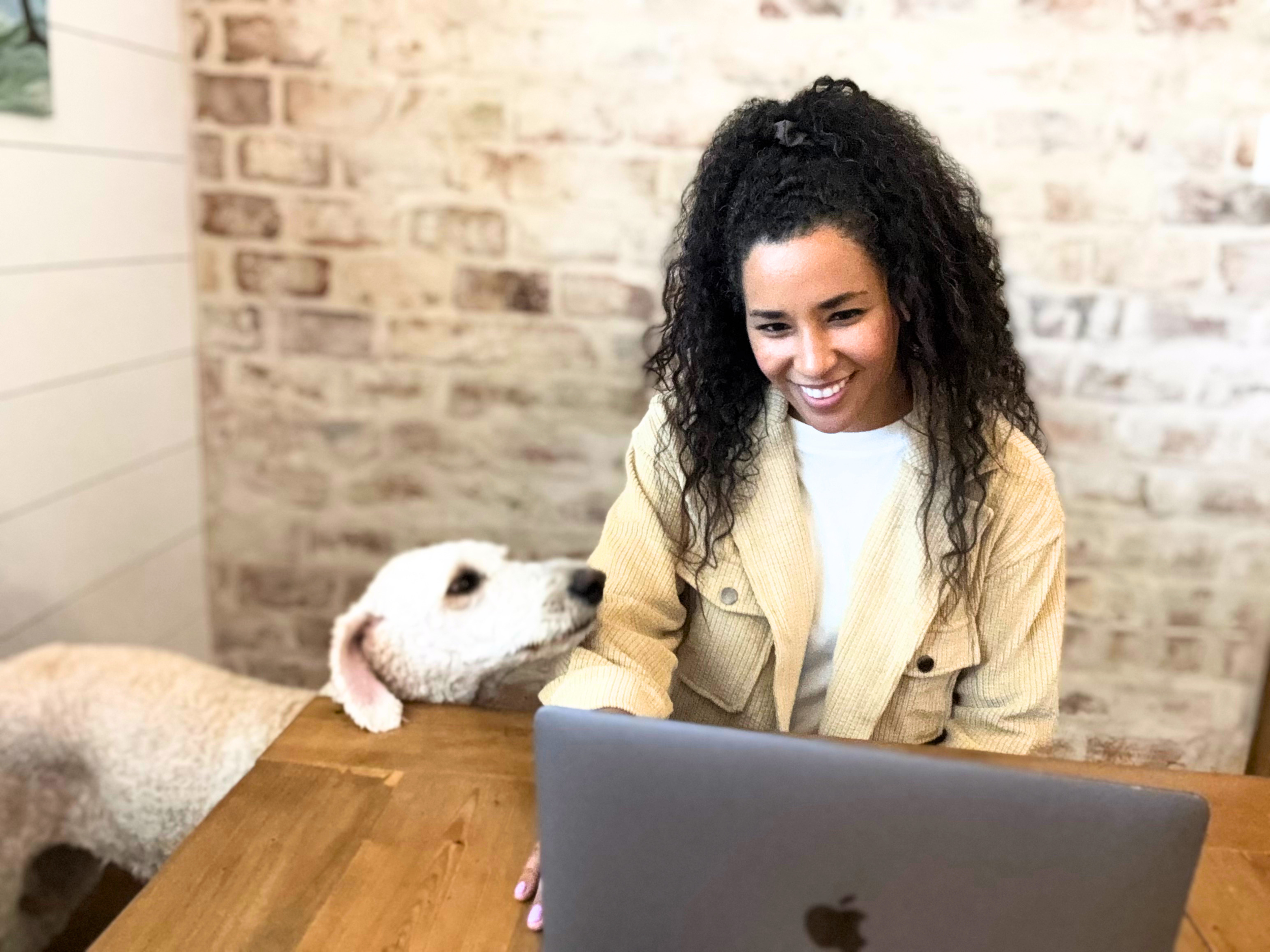 A woman with curly dark hair and a hair clip, wearing a yellow jacket, smiling while looking at her laptop, sitting at a wooden table in a room with brick walls and a white wall.