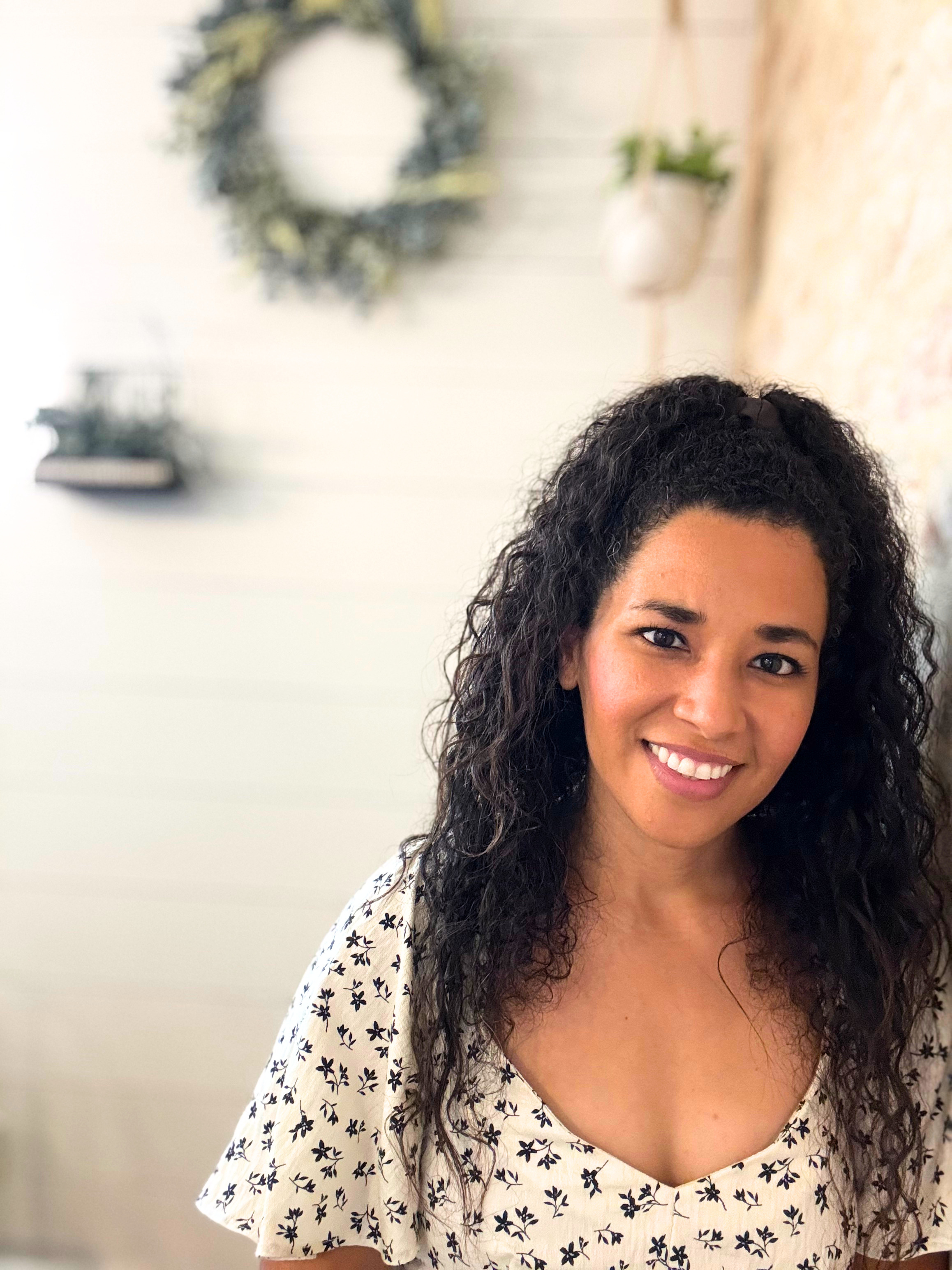 Close-up of a woman smiling with dark, curly hair wearing a white blouse with black floral pattern, in front of a white wall with a decorative wreath and a vase with green leaves in the background.