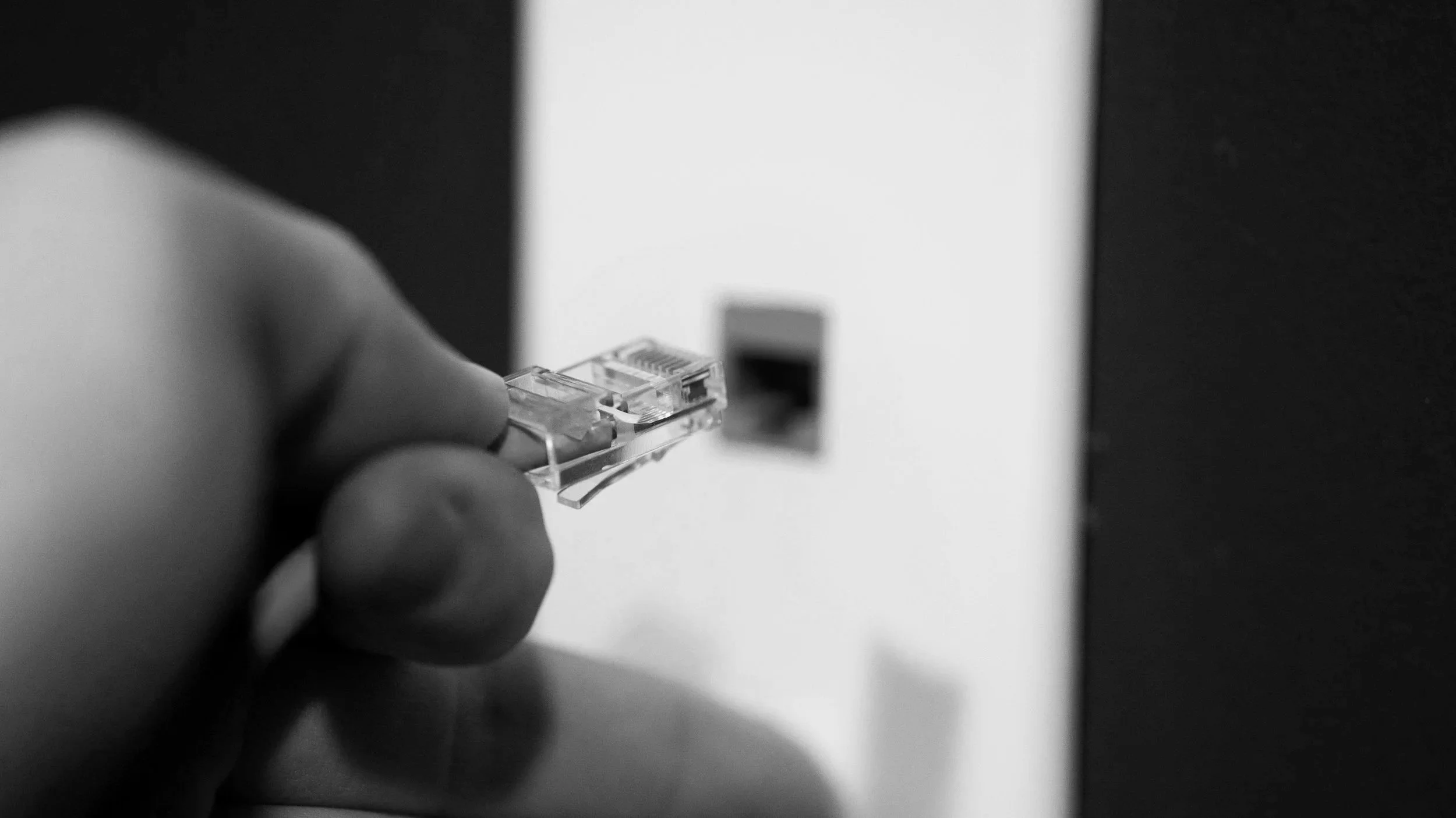 Close-up of a person's hand holding a network cable near an Ethernet port on a white surface, with a dark background.