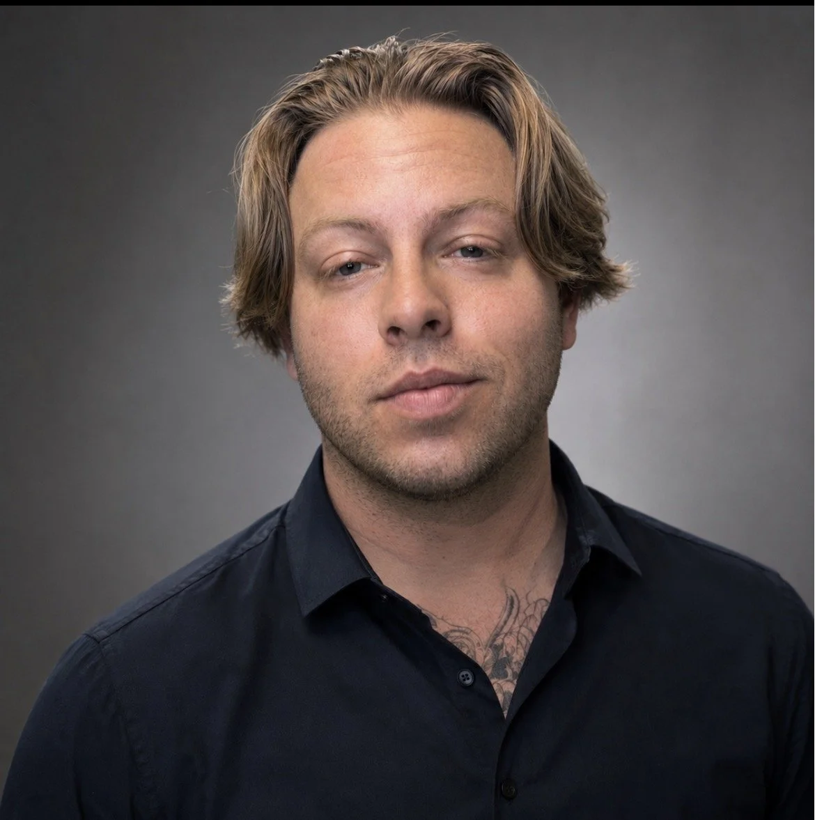 A young man with shoulder-length light brown hair, light skin, and a slight beard, wearing a black button-up shirt, posing against a plain gray background.