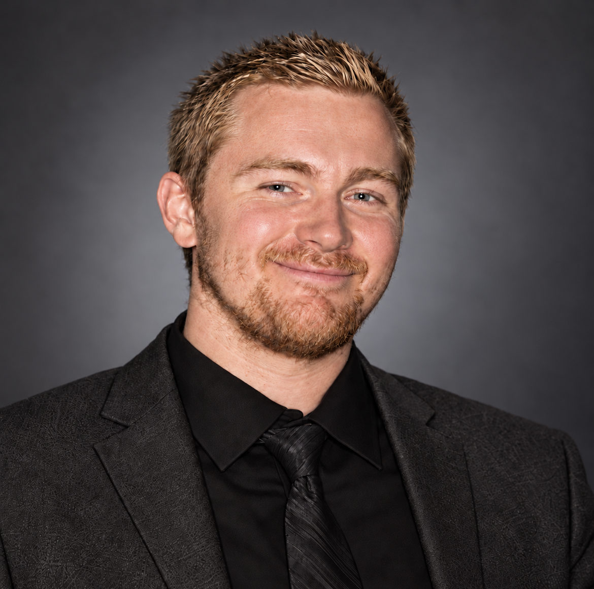 Portrait of a young man with reddish-blonde hair and beard, wearing a black suit, black shirt, and black tie, smiling against a dark gray background.