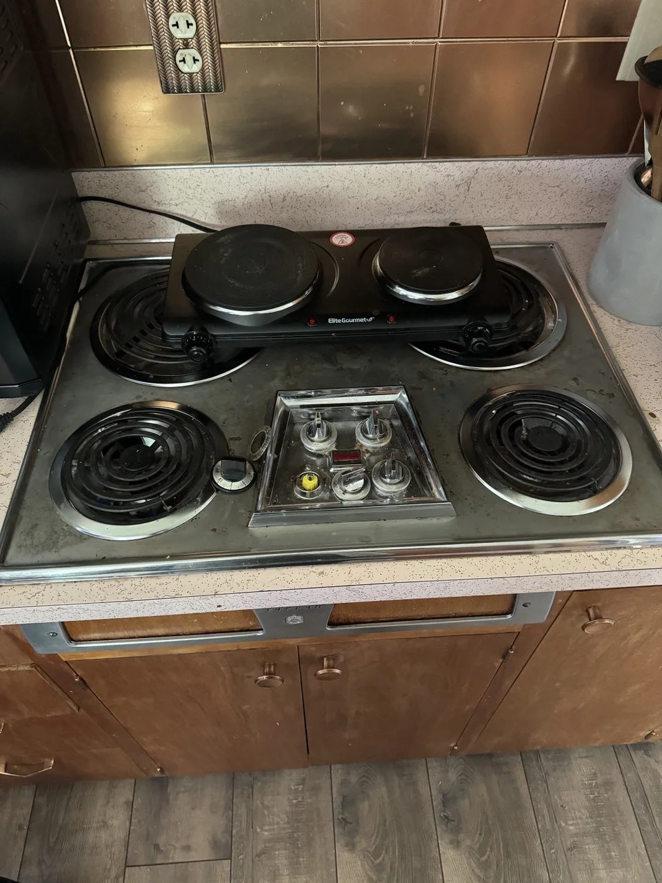 A four-burner electric stove with an additional two-burner hot plate on top, situated in a kitchen with wooden cabinets and a tiled backsplash.