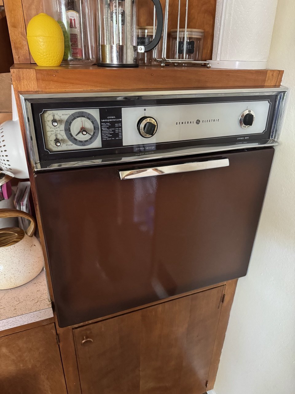 Vintage General Electric oven with a dial control panel, mounted on a wooden cabinet, with kitchen items on the countertop above, including a yellow pineapple-shaped container, a glass container, a coffee jar, and a stainless steel kettle.