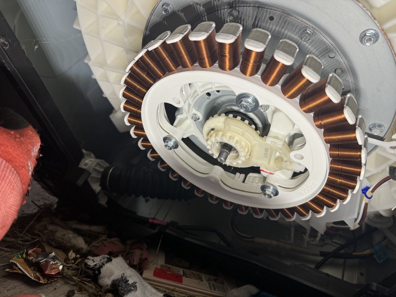 Close-up of an electric motor inside a washing machine, showing the stator with copper wire windings surrounding a central gear and bearing, with some debris at the bottom.