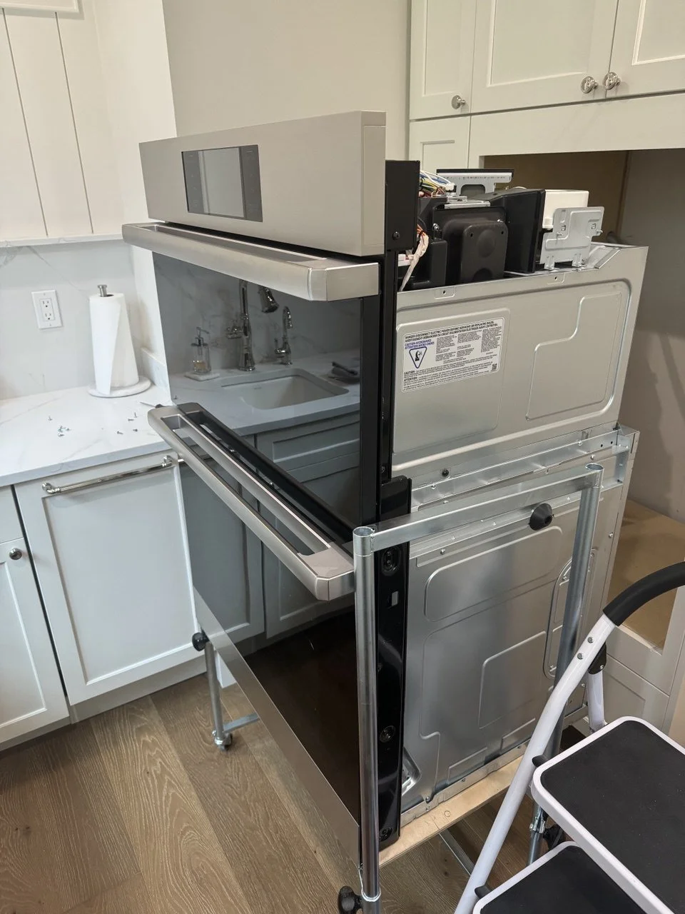 Disassembled kitchen dishwasher next to a white cabinet and sink, with a step stool nearby.