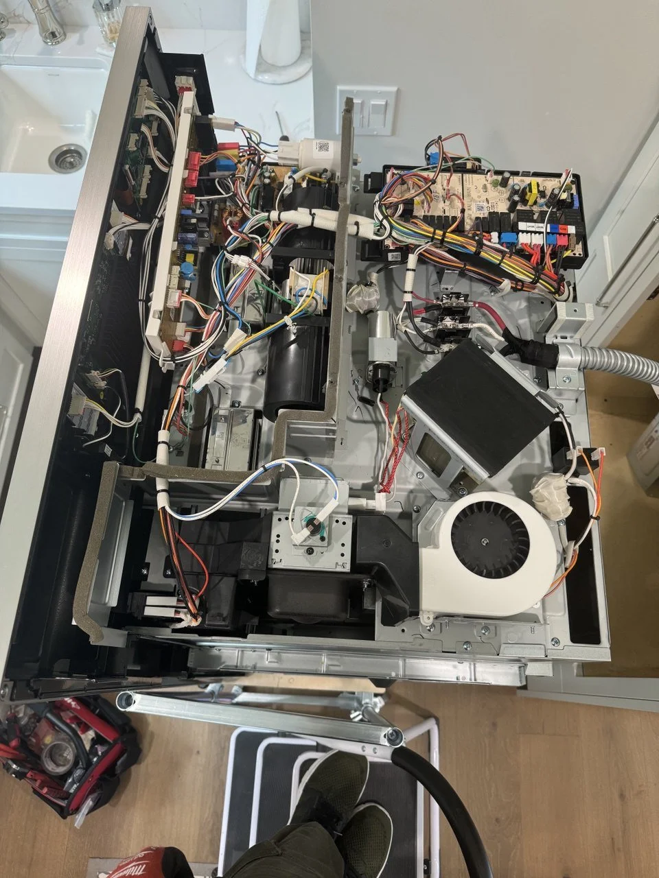 The image shows the open internal components of a countertop kitchen appliance, likely a coffee machine, revealing various wires, circuit boards, motors, and mechanical parts.