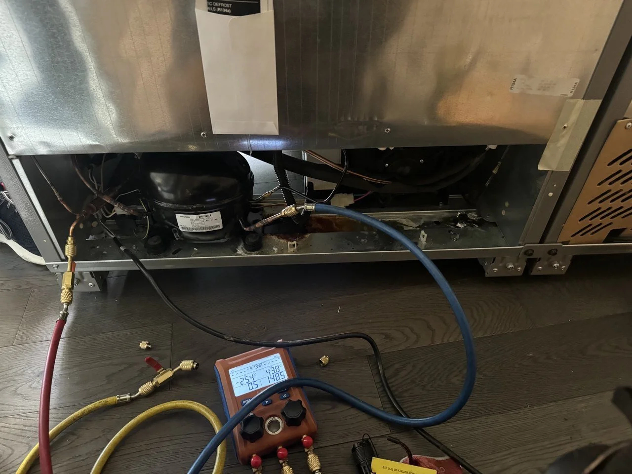 The interior of a refrigerator showing the compressor and related components, with electronic tools and hoses connected for testing or repair.