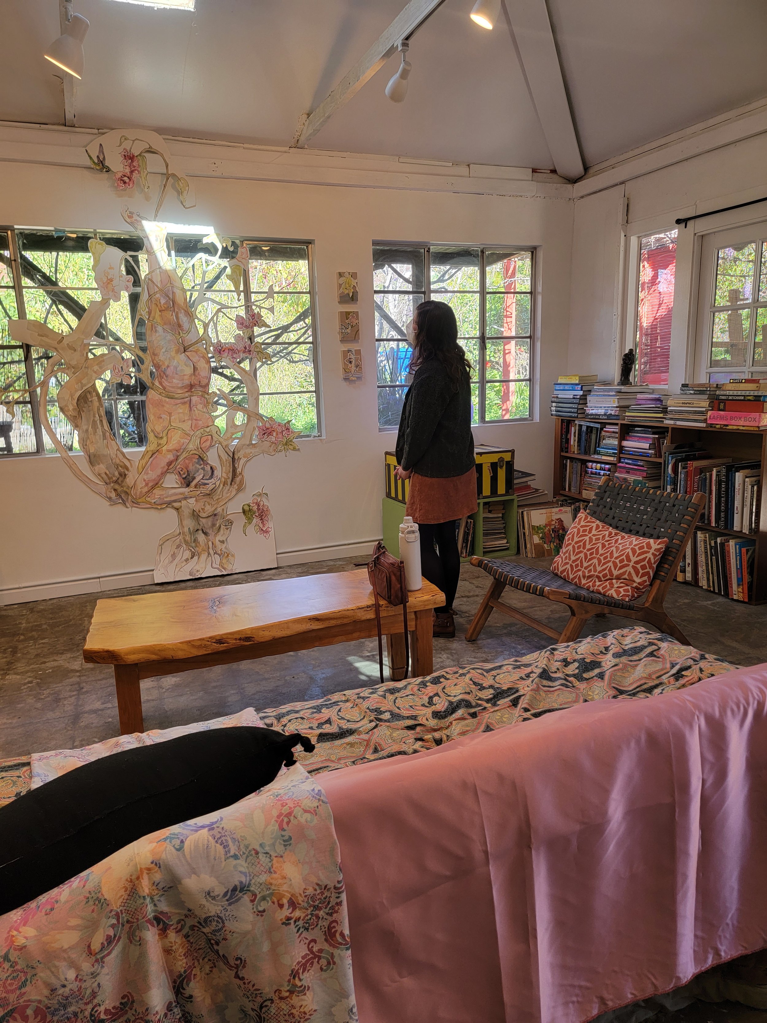 Person standing in an art gallery, looking at a large painting of a tree with pink flowers on the wall. The room has white walls, large windows, a bookshelf filled with books, a wooden bench, and a patterned sofa with a pink cover and pillow.