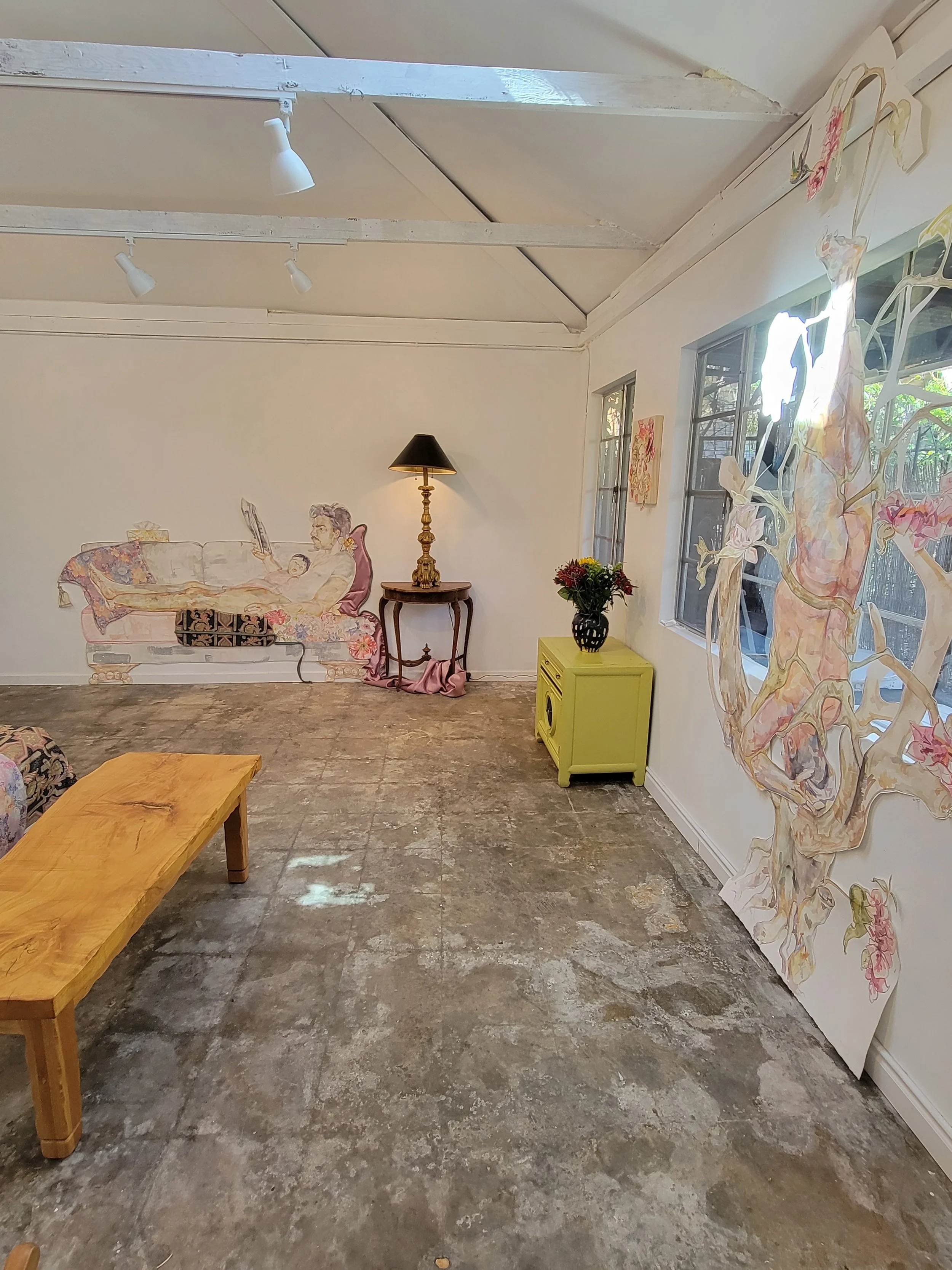 Interior of an art gallery with a mural of a woman reading on a sofa, a yellow side table with a vase of flowers, a wooden coffee table, a wall with floral paper art, and windows letting in natural light.