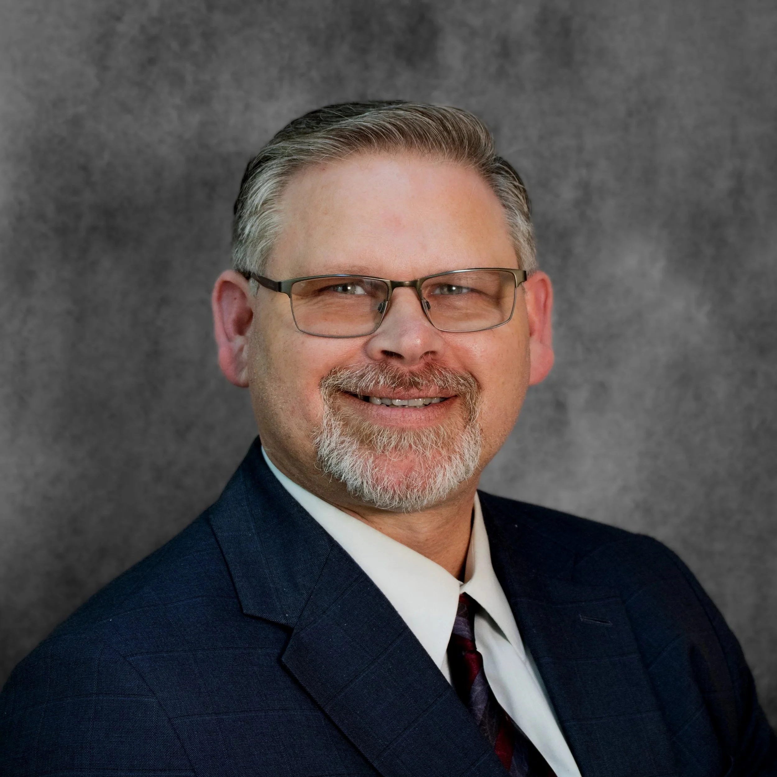 Close-up professional portrait of a middle-aged man with glasses, gray hair, and a beard, smiling, wearing a dark suit, white shirt, and golf tie, against a gray background.