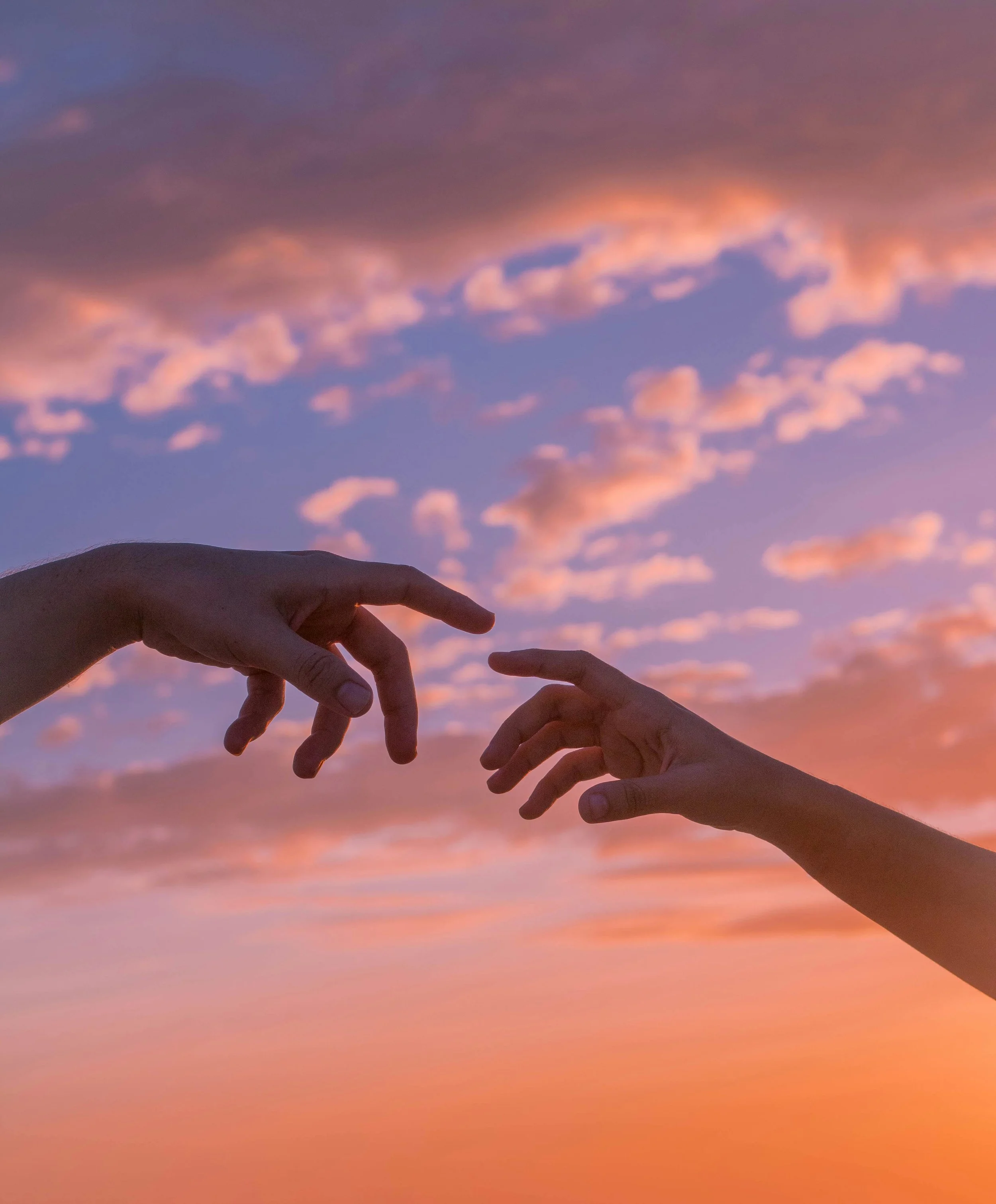 Two hands reaching towards each other against a colorful sunset sky with scattered clouds.