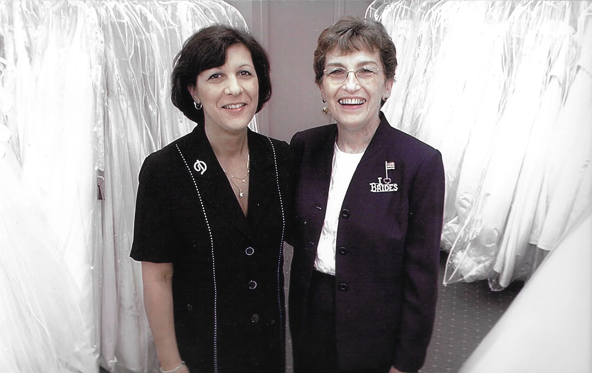 Two women standing together, smiling in front of bridal dresses on display, one wearing a dark blazer with a 'Bride' pin and the others in dark and pearl accents.