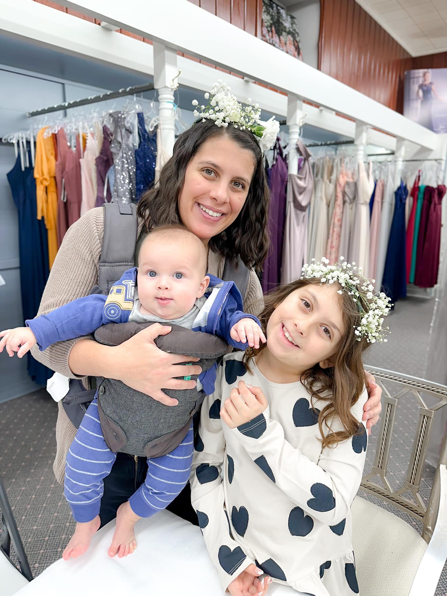 A woman and two children with floral crowns in a dress shop, surrounded by colorful dresses on racks.