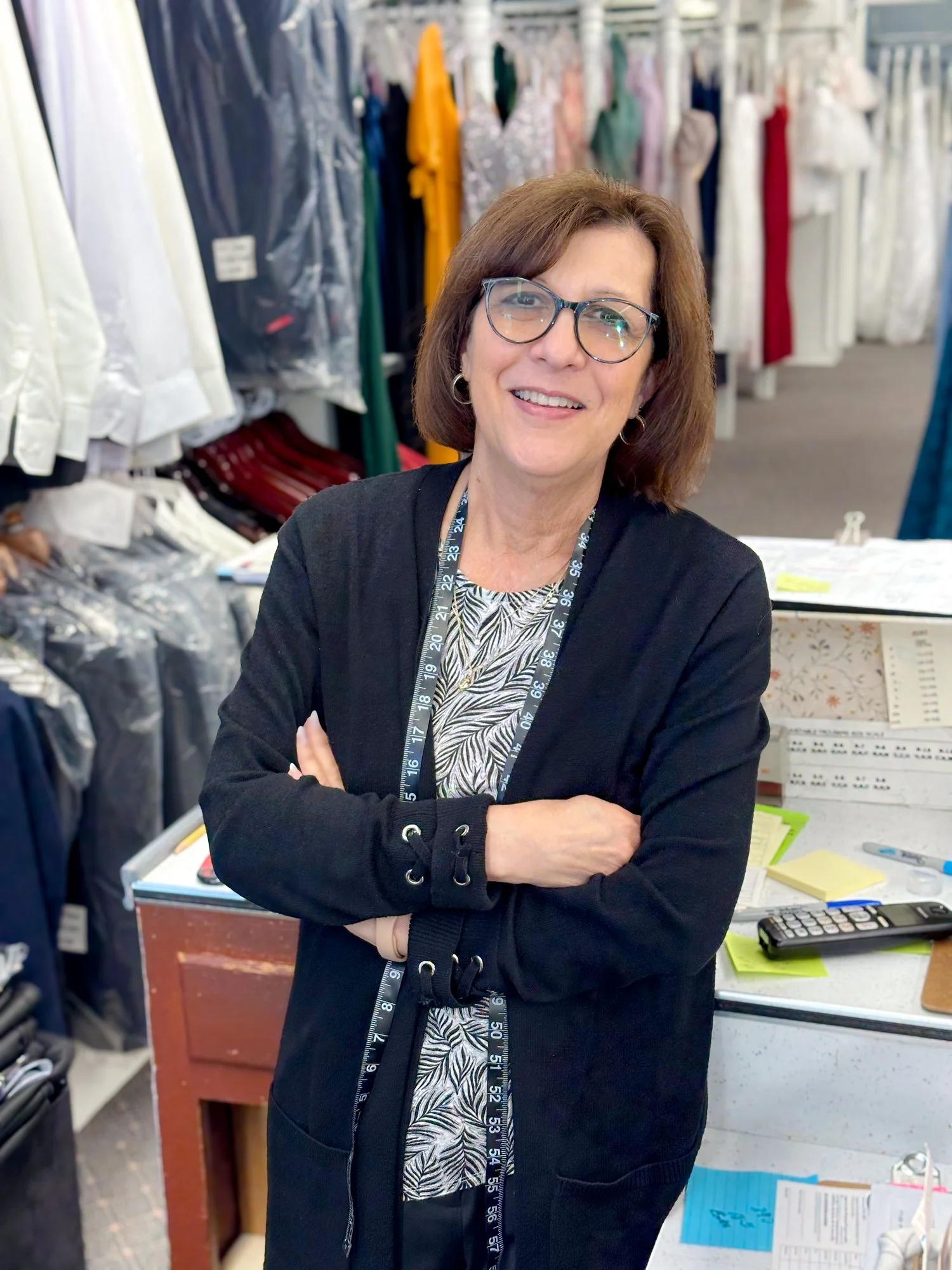 A woman with glasses, wearing a black cardigan, standing with arms crossed, smiling in a fabric store with hanging fabric bolts and a desk with papers and a calculator in the background.