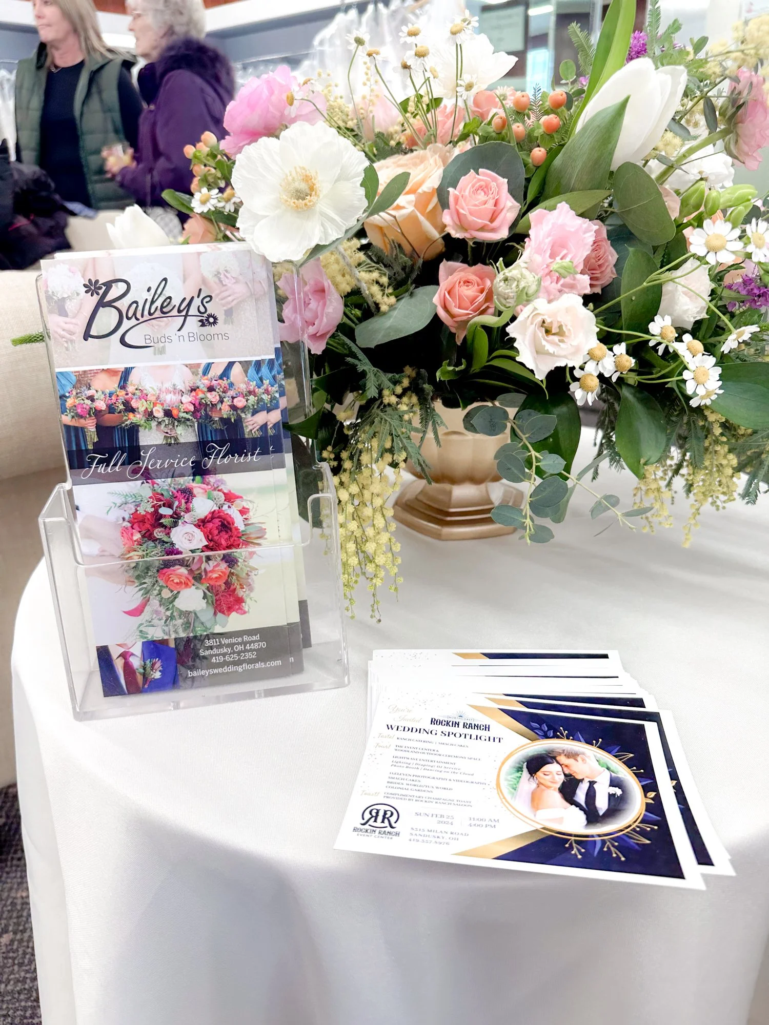 A large bouquet of pink and white flowers on a round table with a wedding flyer and business card holder nearby, and blurred people in the background.