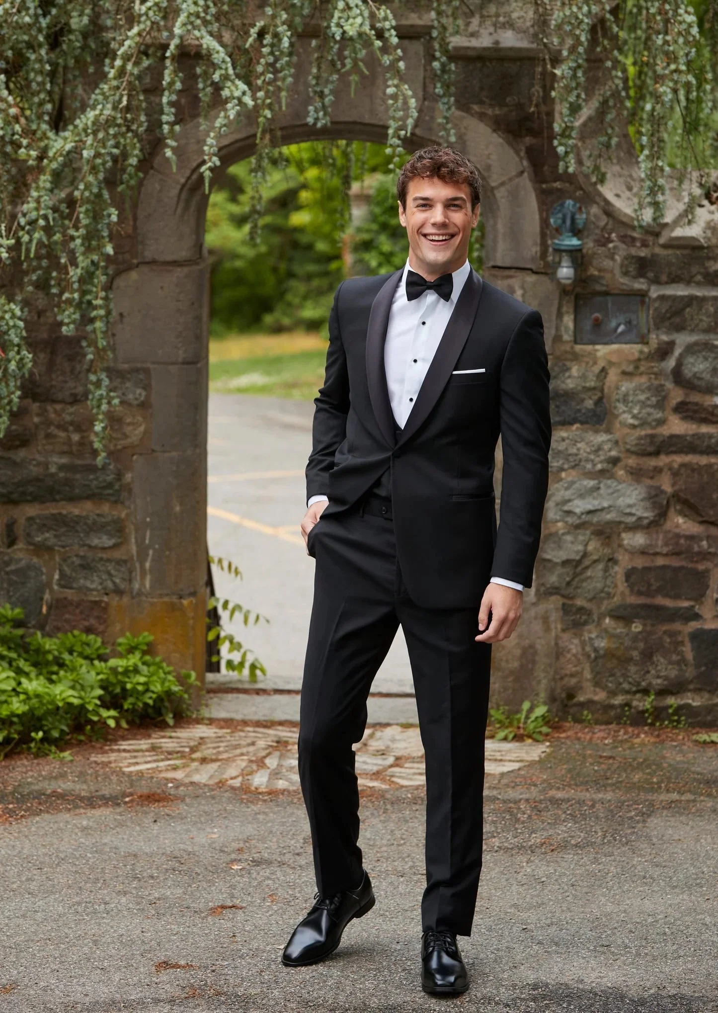 Young man in a black tuxedo with a bow tie, standing outdoors in front of a stone archway decorated with hanging greenery, smiling at the camera.