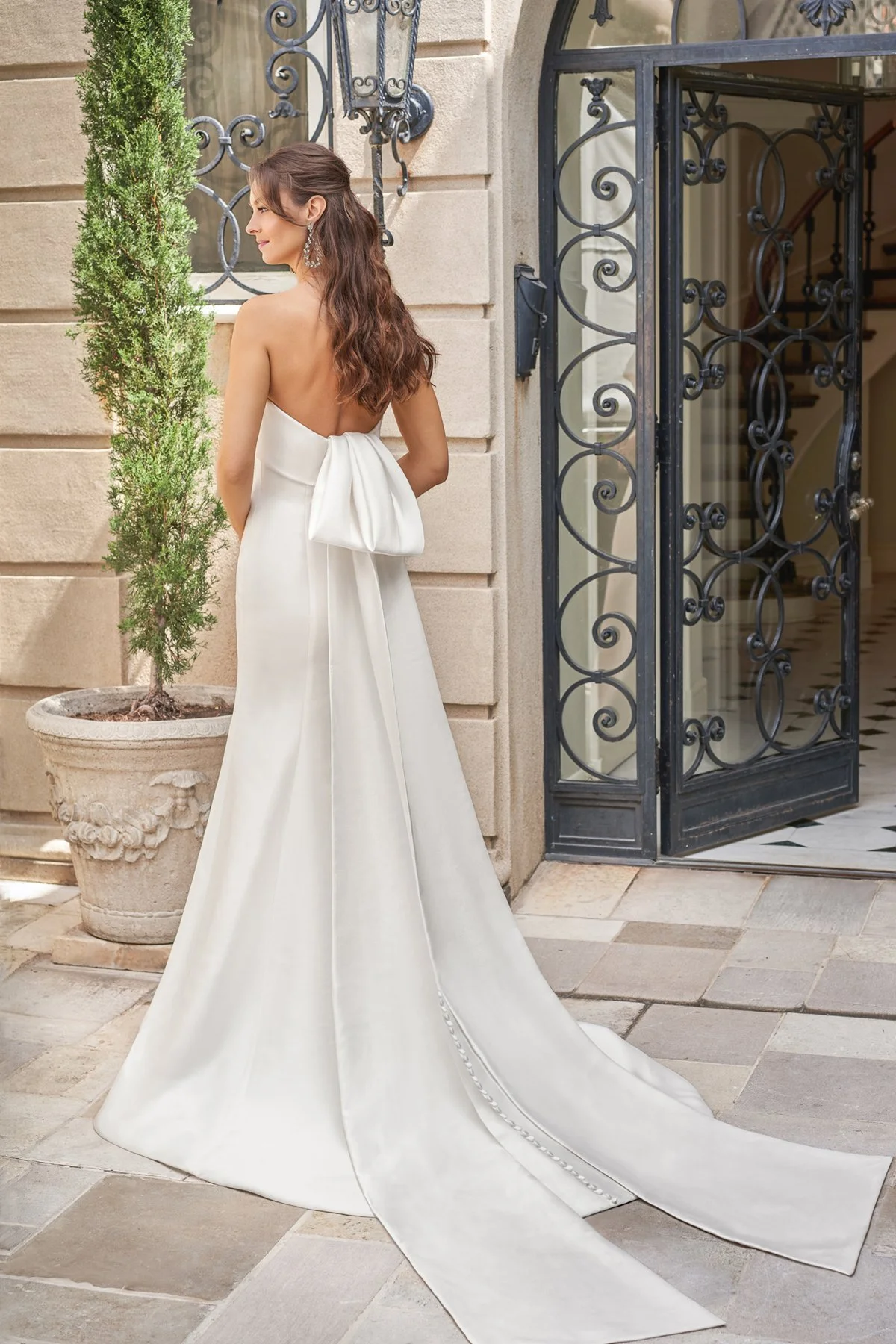 A woman in a strapless white wedding dress with a large bow at the back, standing outside a building with an ornate black iron gate, next to a potted plant.