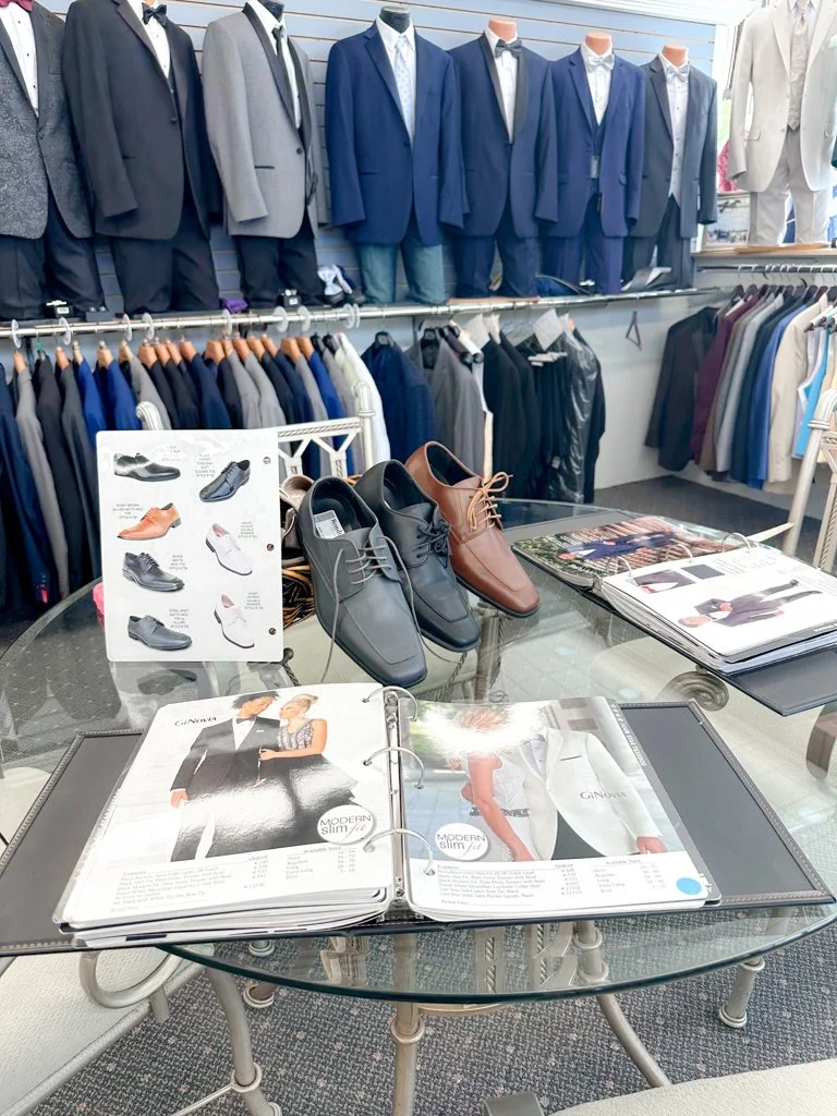 Display of men's formal suits on mannequins, leather dress shoes, and suit catalogs in a clothing store.