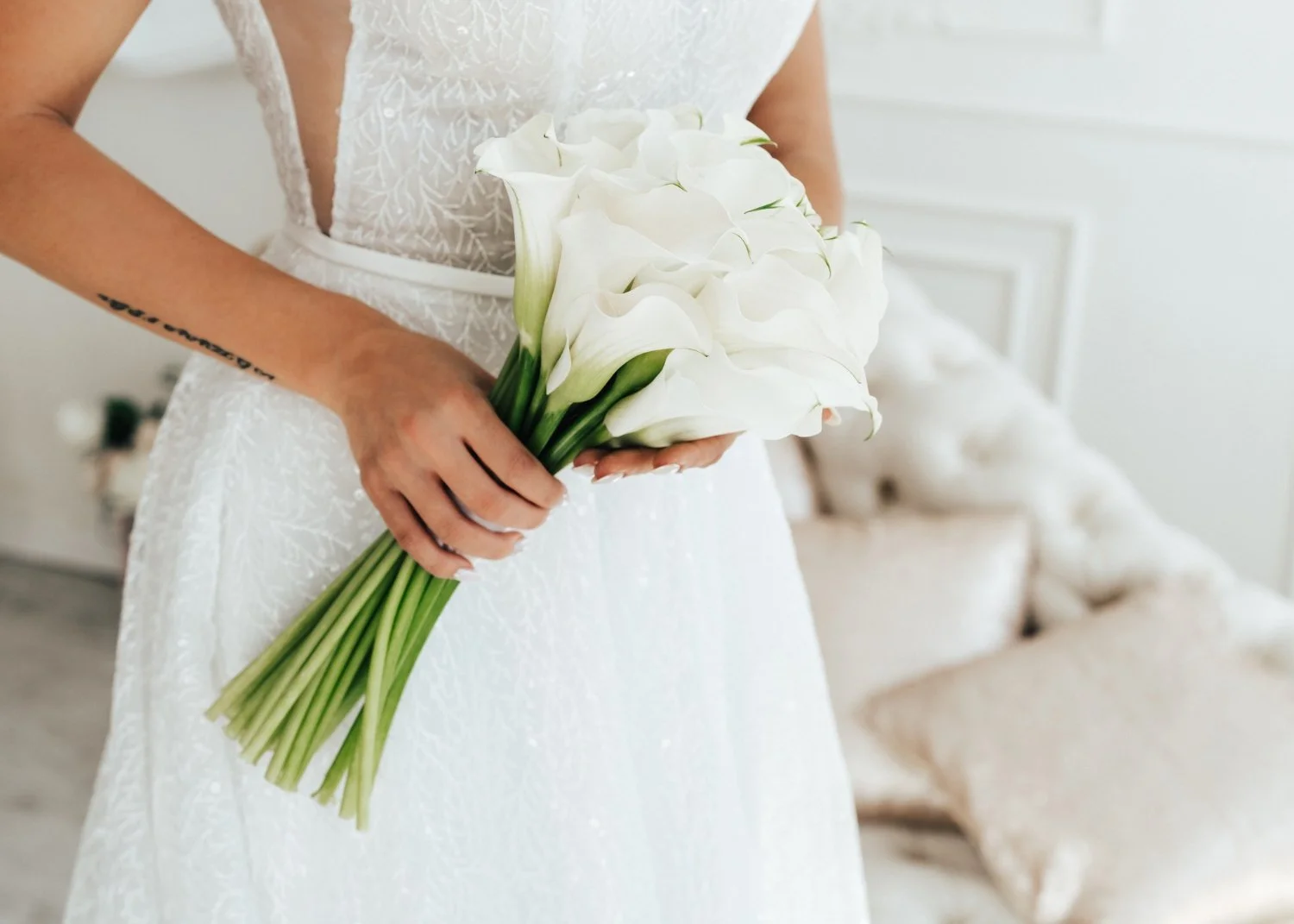 A woman in a white lace dress holding a bouquet of white calla lilies in a bright, elegant room with cream-colored furniture.