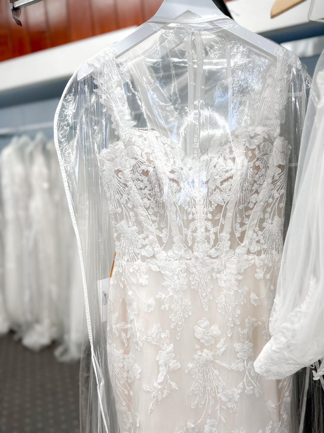 A white wedding dress with lace embroidery is hanging on a clothing rack, covered with a clear plastic garment bag.