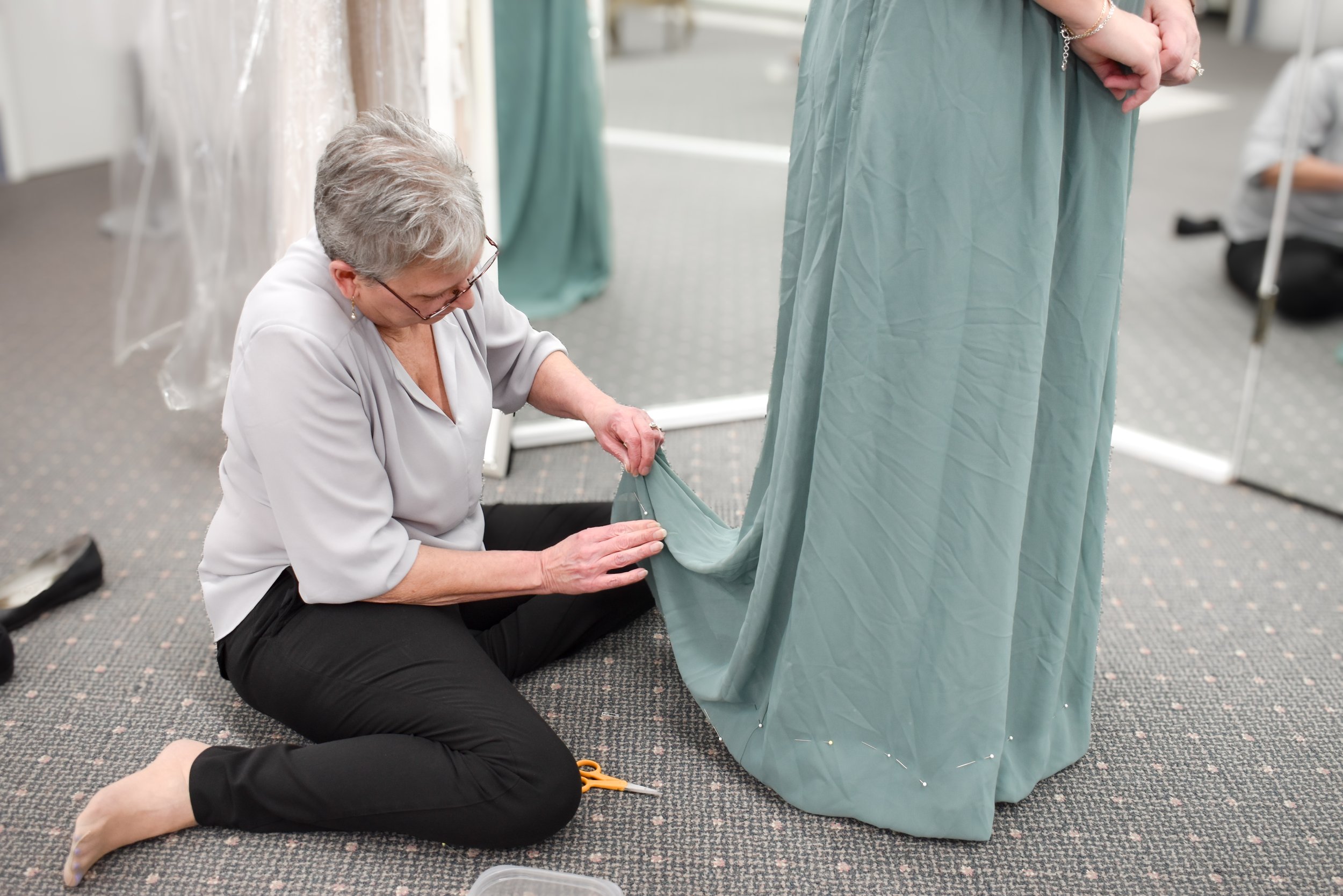 An older woman is sitting on the floor measuring and pinning a piece of fabric or costume, with a pair of scissors nearby, while working on a costume or dress in a room that looks like a dressing or costume area.