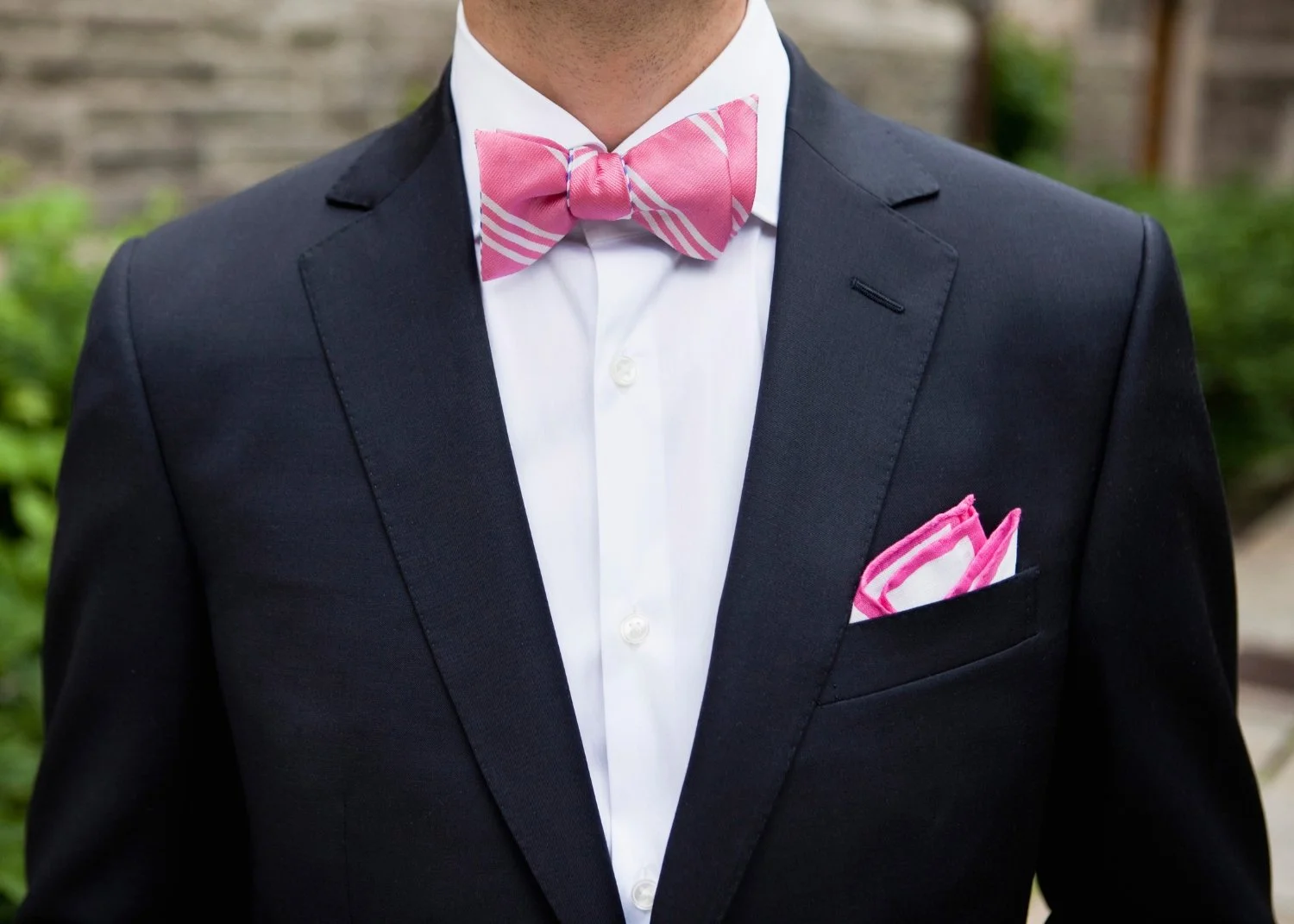 Man in navy blue suit with pink and white striped bow tie and pocket square, white dress shirt, outdoor setting with greenery and brick wall.