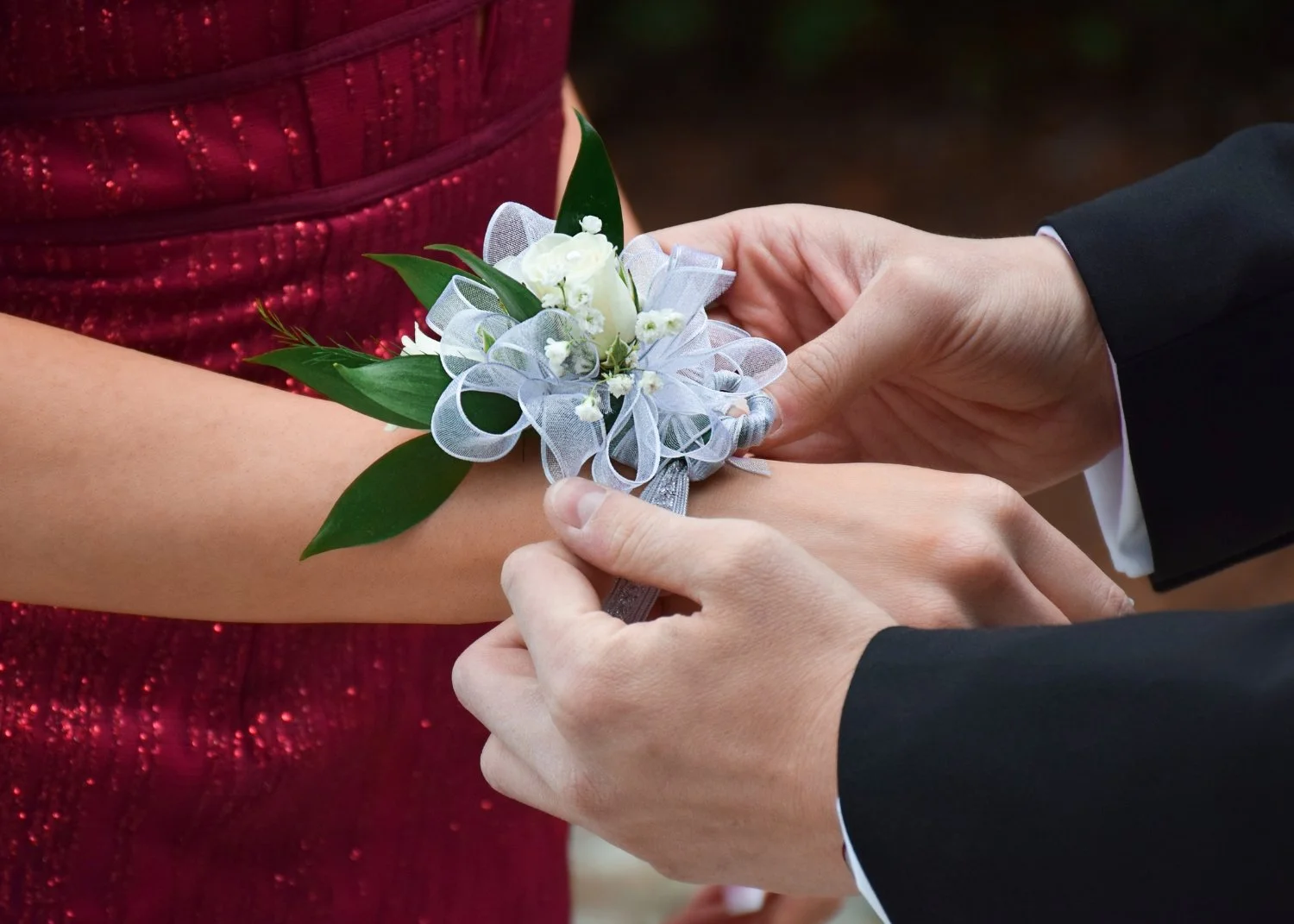 Close-up of a man placing a white corsage with white flowers and ribbons on a woman's wrist during a formal event.