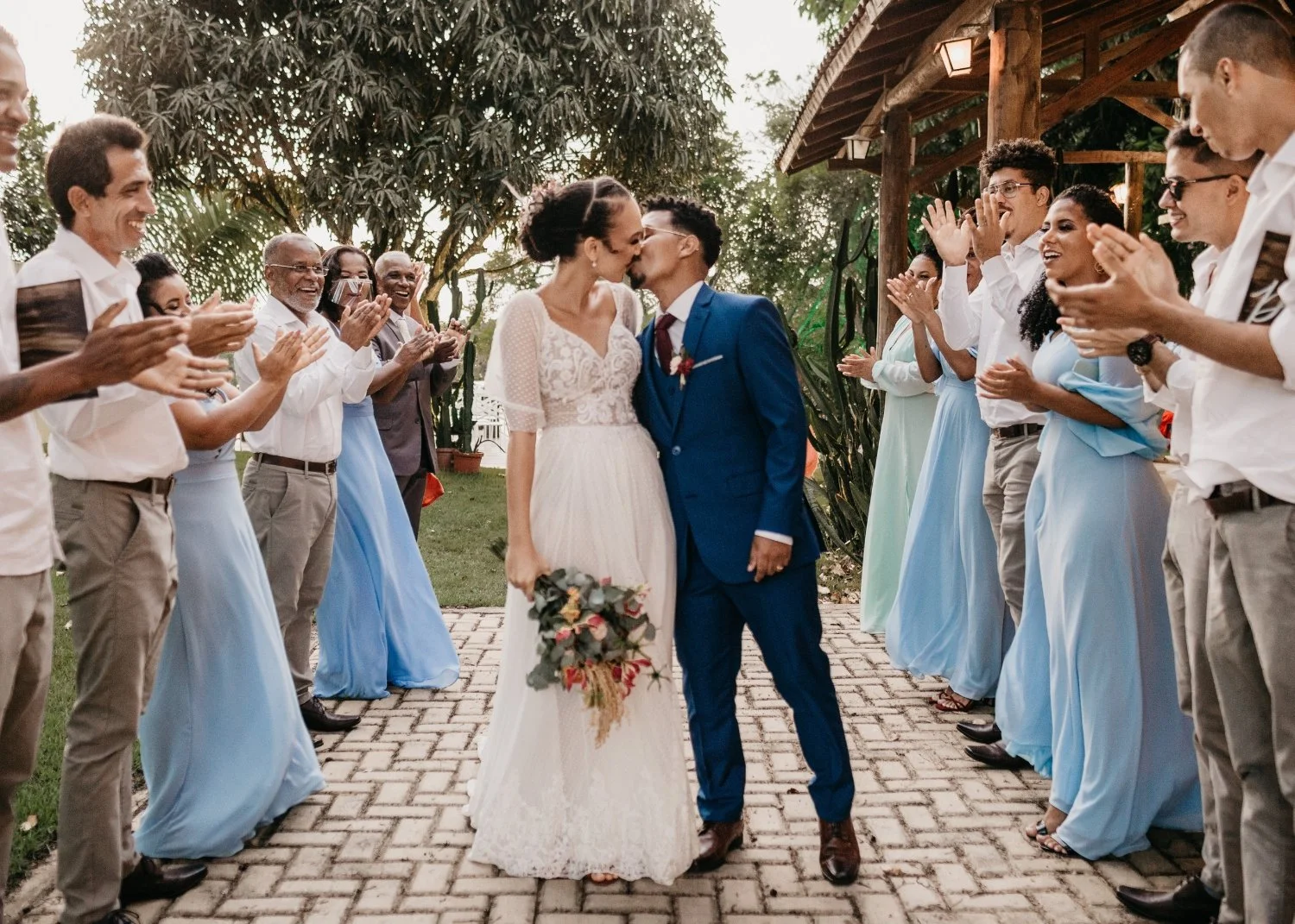 Couple in wedding attire kissing under a canopy with wedding guests clapping and smiling on each side.