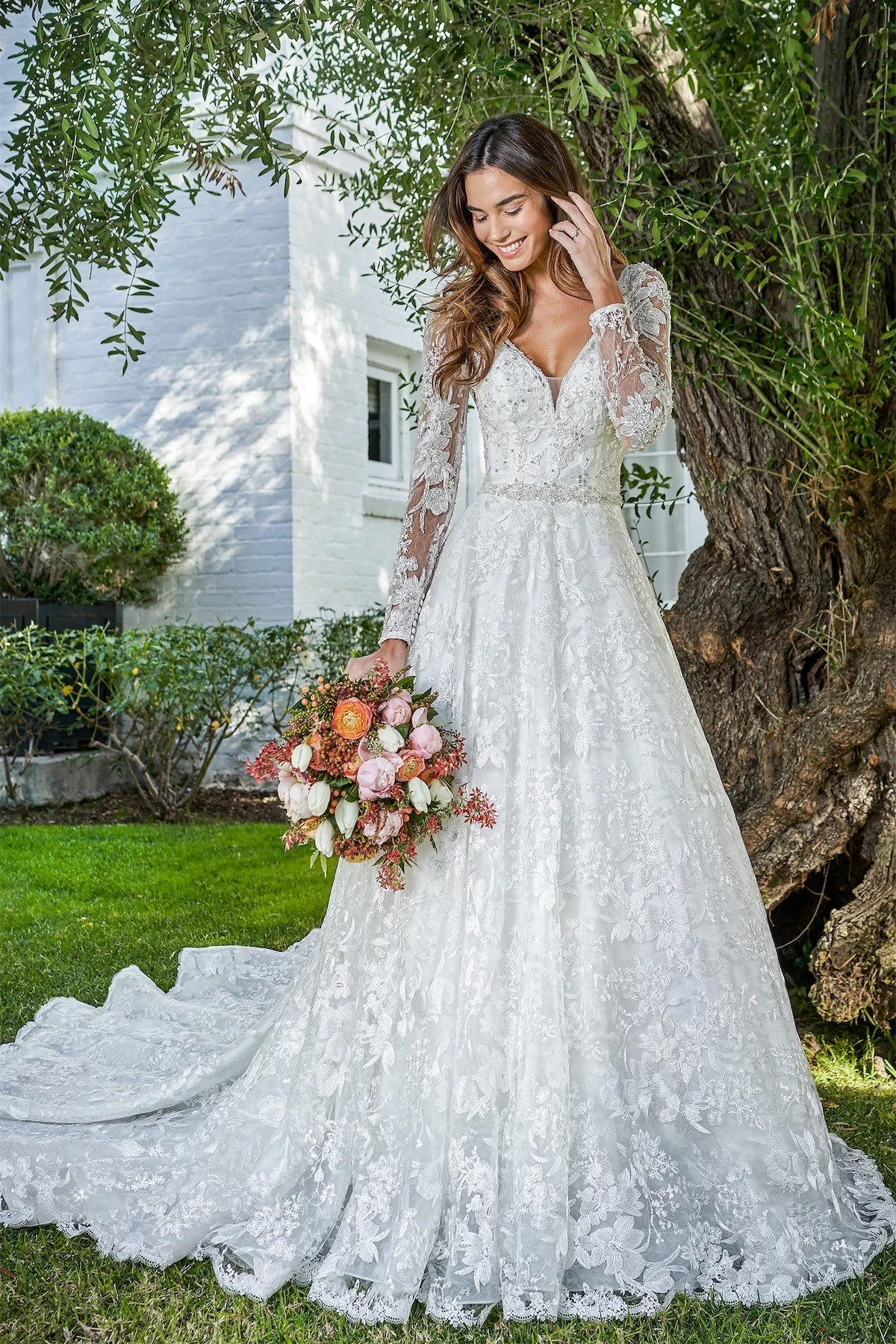 A woman in a white wedding dress with long lace sleeves stands outdoors near a tree, holding a pink and orange bouquet, smiling.