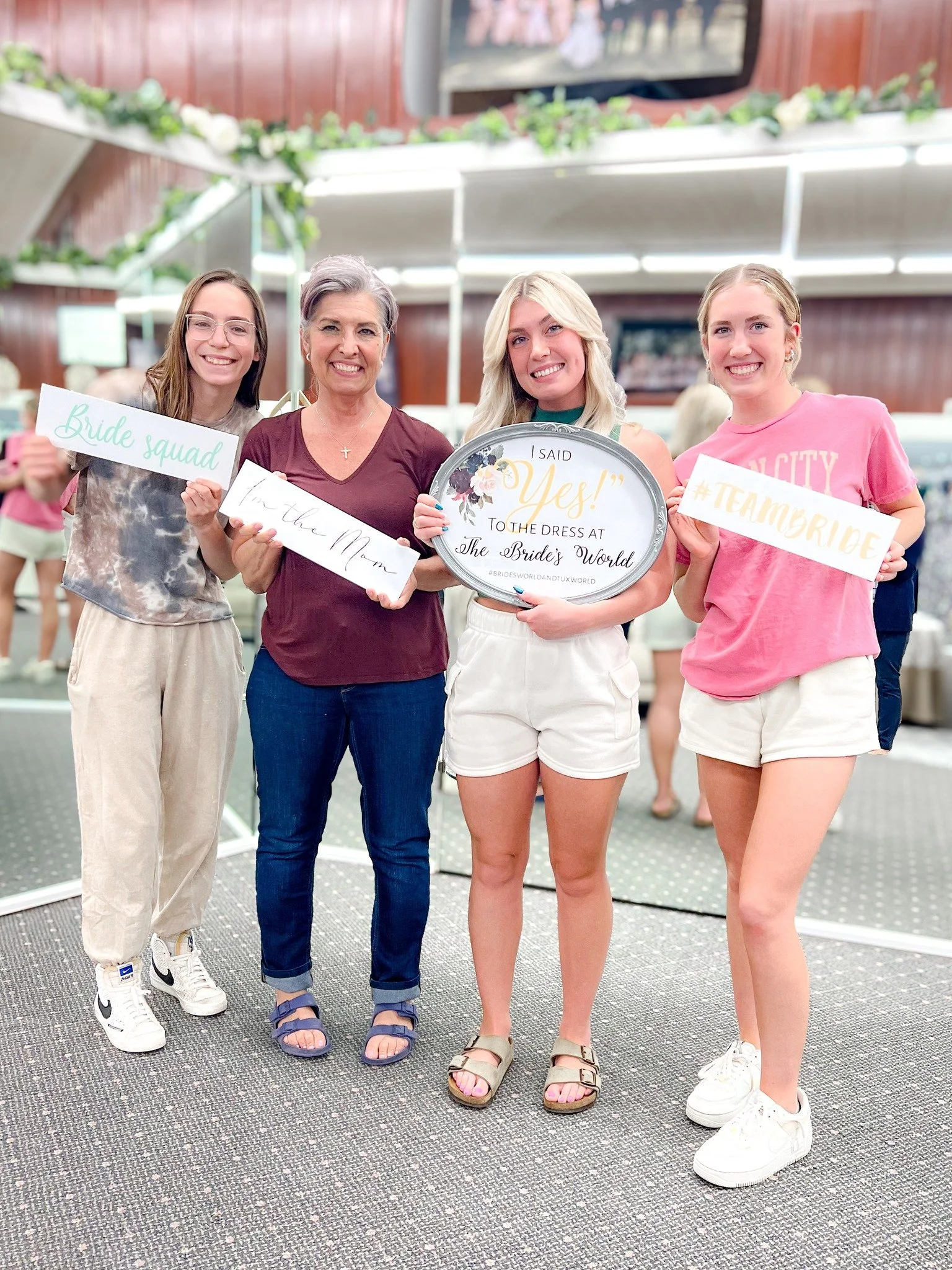 Group of four women at a bridal shop, with one holding a sign that says "Yes! I said yes! to the dress at the bride's world," and others holding signs that say "bride squad," "till the mom," and "#TeamBride."