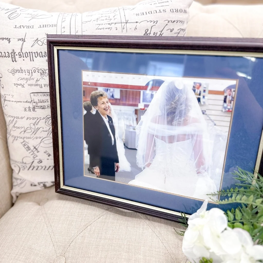 A framed wedding photograph showing an older woman smiling at a bride, who is wearing a wedding dress and veil, inside a store or display area.