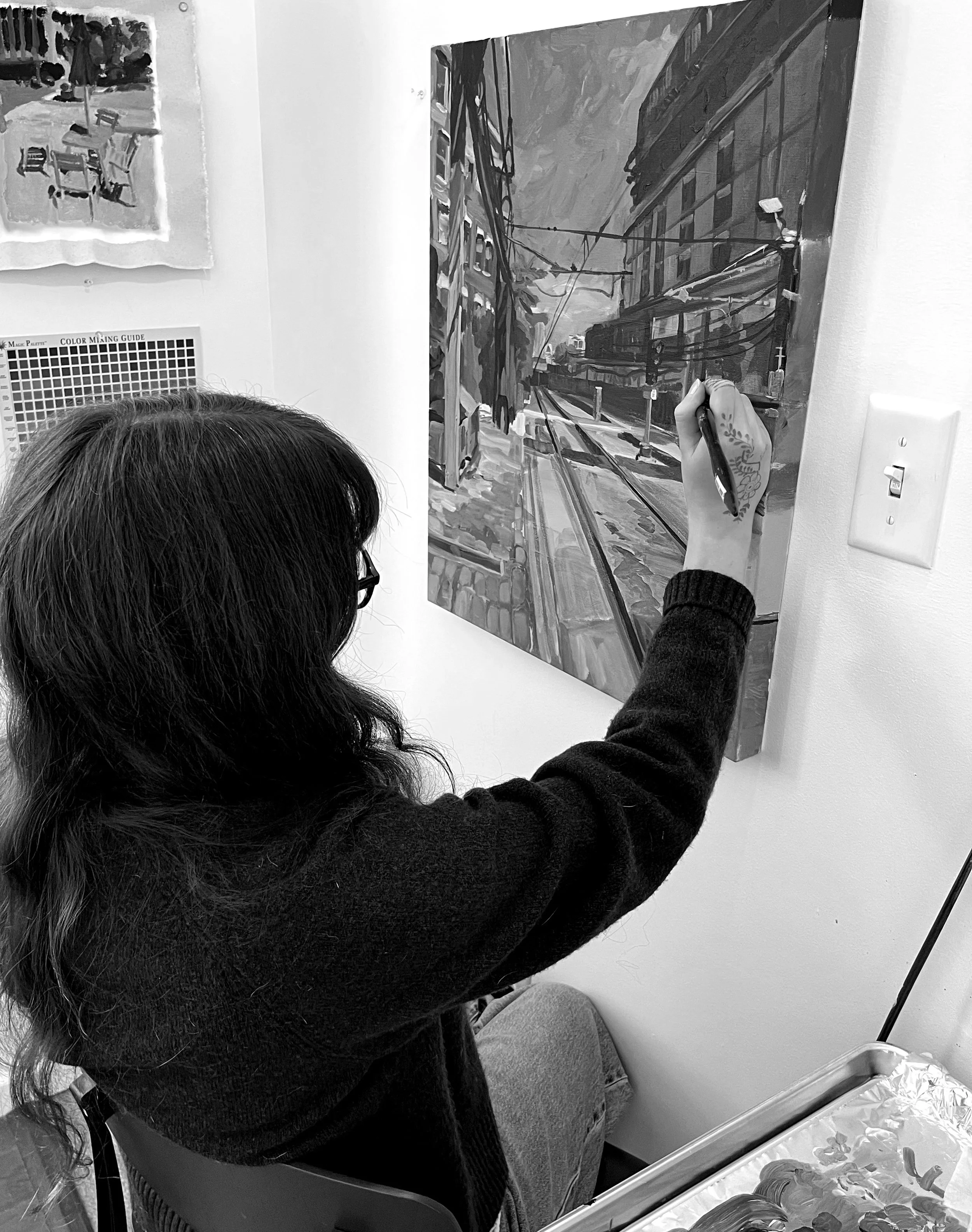 Picture of the artist sitting in a chair and painting, facing away from the camera
