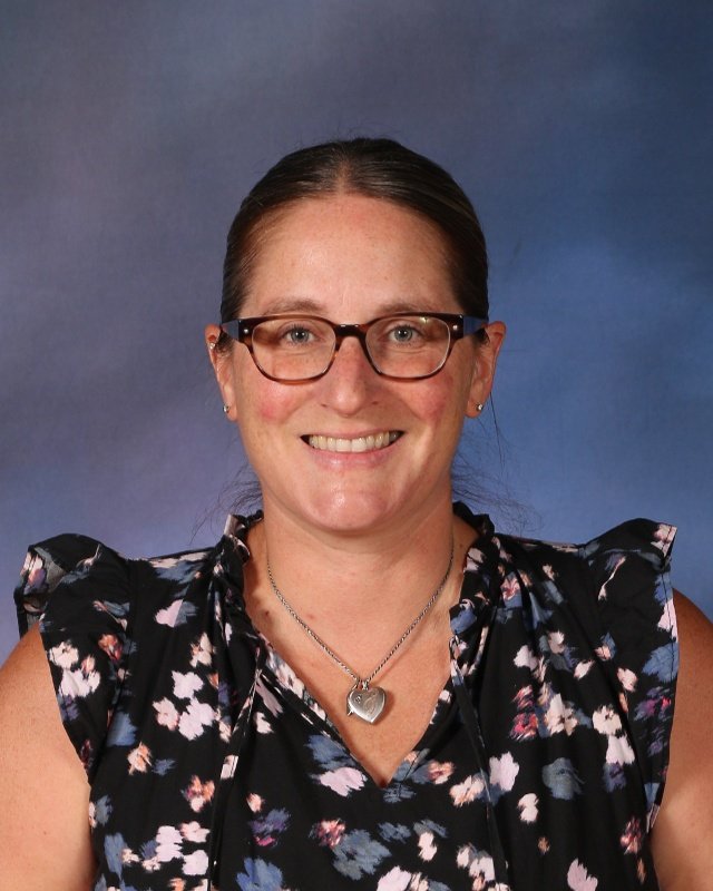 A smiling woman with brown hair, glasses, and earrings, wearing a floral top with ruffled sleeves and a heart-shaped pendant necklace, against a blue background.