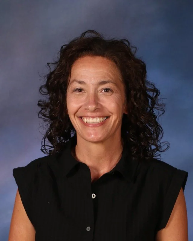 A woman with curly brown hair wearing a black sleeveless collared shirt, smiling at the camera against a blue gradient background.